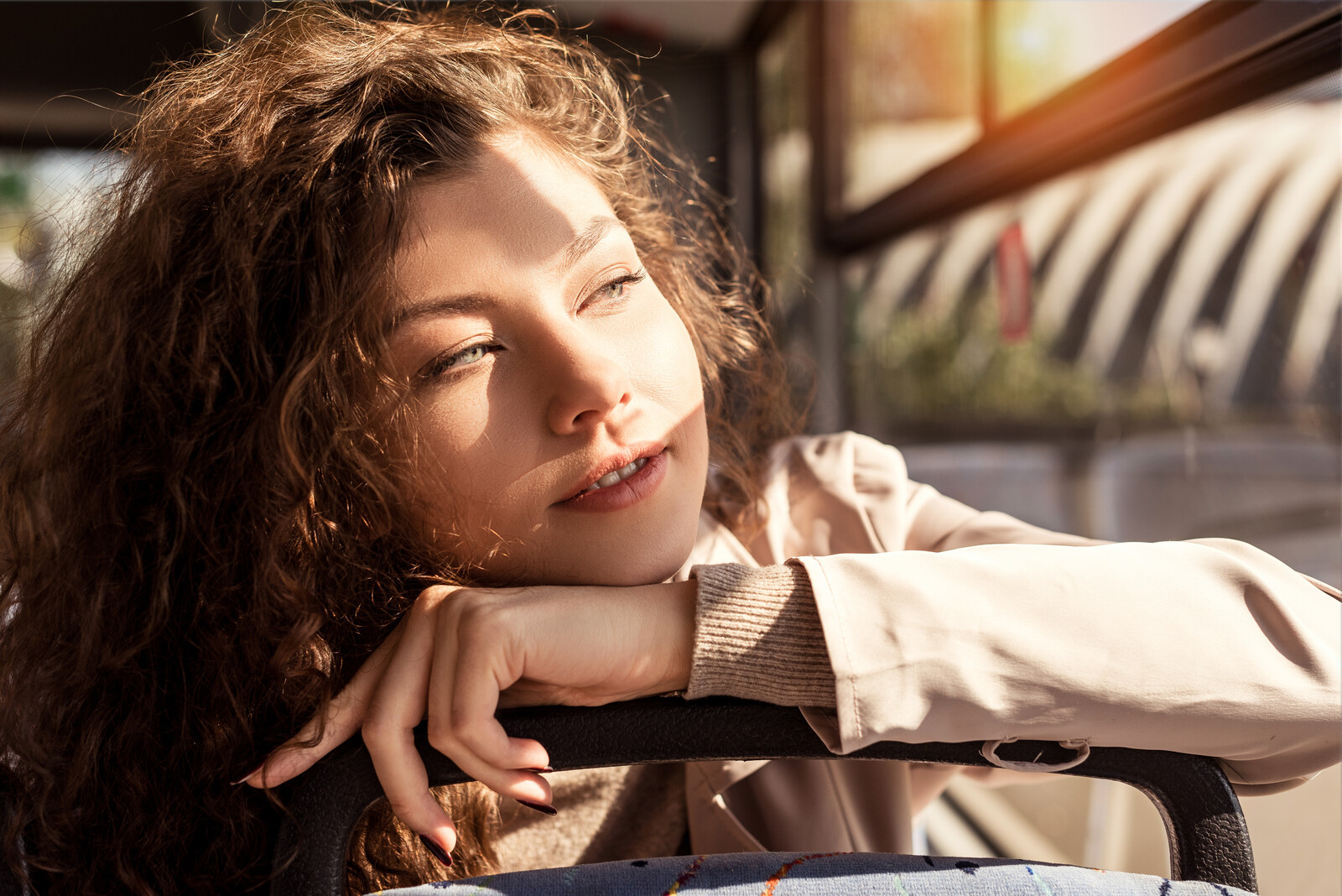 A woman with curly brown hair rests her chin on her hands, which are placed on the back of a bus seat. Sunlight streams through the window, casting a warm glow on her face as she gazes thoughtfully outside.