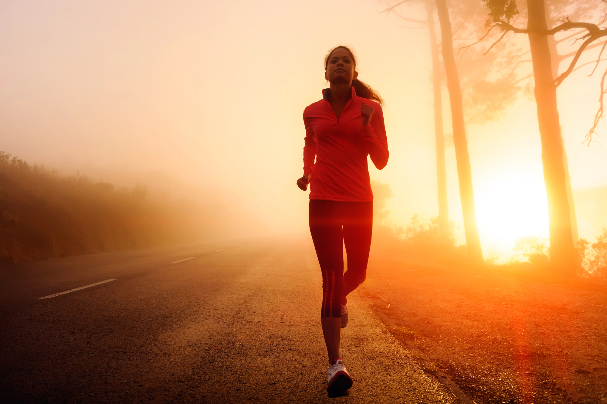 A person is jogging on a road at dawn or dusk, wearing a red long-sleeved top, black leggings, and running shoes. The sun is rising or setting in the background, casting an orange glow and creating a misty atmosphere among the trees along the roadside.