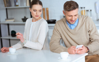 A woman in a white top looks over at a man in a beige sweater who is smiling while using his smartphone. They are sitting at a white table with coffee cups in an indoor setting. The woman appears curious or possibly suspicious.