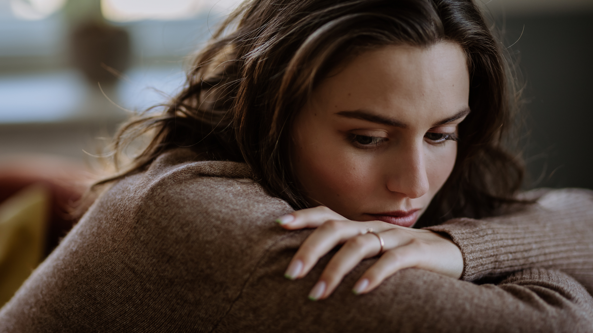 A woman with long brown hair is resting her head on her folded arms, looking contemplative or sad. She is wearing a brown sweater, and her expression suggests deep thought or concern. The background is blurred, focusing attention on her face.