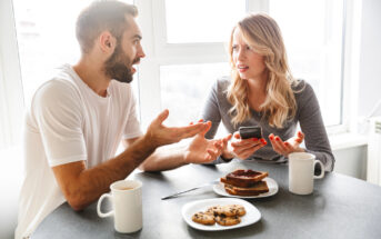 A man and woman are sitting at a table near a window, having a conversation. The man gestures with his hands, while the woman looks at him, holding a phone. On the table are two mugs, a plate with cookies, and a plate with toast and jam.