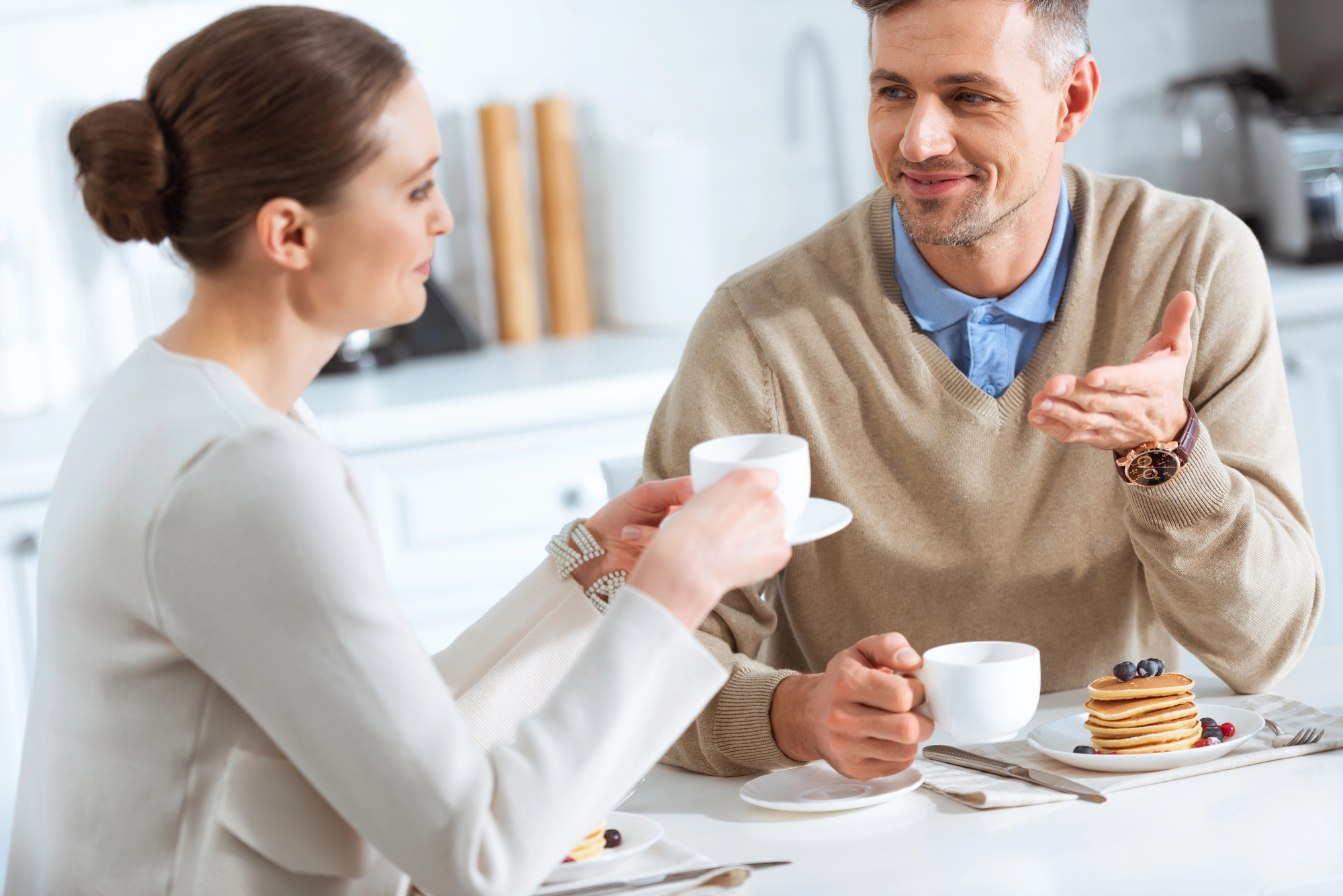 A man and woman sit at a table in a bright kitchen, enjoying coffee and pancakes. The man gestures with his hand while talking, and the woman holds a coffee cup, attentively listening. Both are casually dressed and appear engaged in conversation.