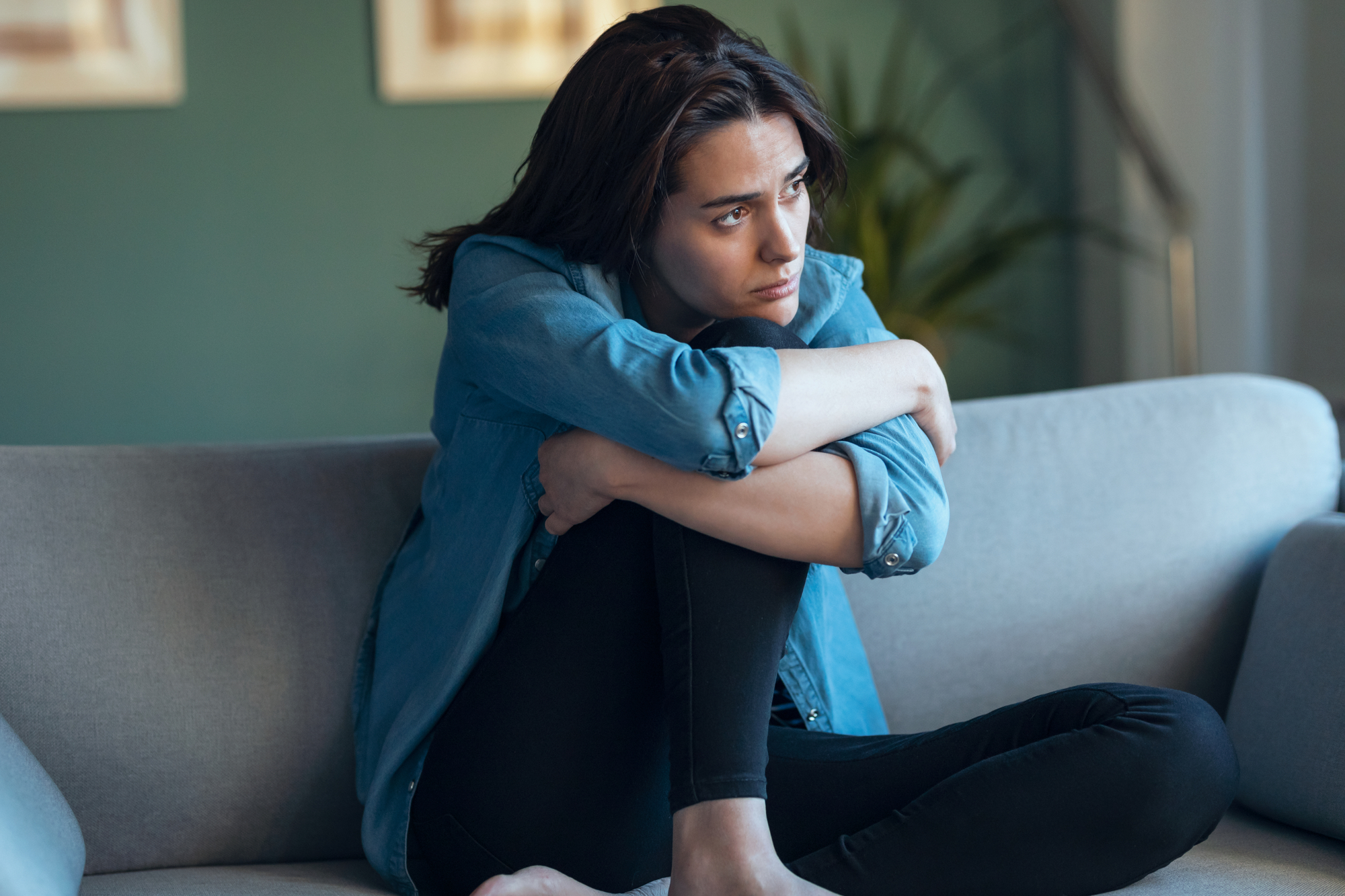 A pensive person sits on a grey couch, hugging their knees and gazing off into the distance. They have dark hair and wear a light blue denim shirt and black pants. The background includes a green wall and out-of-focus framed pictures.