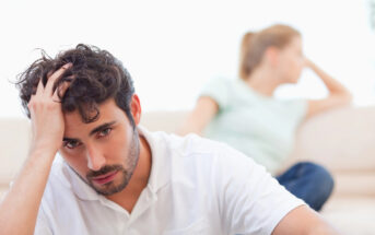 A man in a white shirt sits in the foreground with a distressed expression, holding his head with one hand. In the background, a woman in a light green shirt sits turned away, gazing into the distance. Both appear to be in a tense or thoughtful mood.