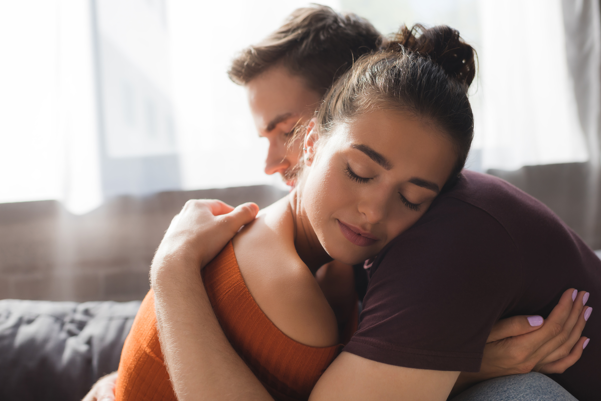 Two people are embracing each other lovingly in a bright room. The person facing the camera has her eyes closed, wearing an orange top. The person facing away has their eyes closed, with their head resting near her shoulder. Both appear calm and content.