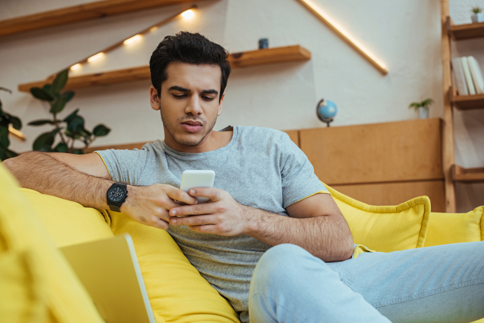A man in a gray t-shirt and light blue jeans is sitting on a bright yellow couch, using a smartphone. He appears to be concentrating or puzzled by what he's seeing on the screen. The background shows wooden shelves with books and decor items.