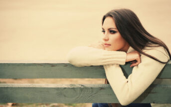 A woman with long brown hair and a cream sweater rests her arms on the back of a weathered wooden bench, gazing thoughtfully into the distance. The background is blurred, suggesting an outdoor setting on a calm day.