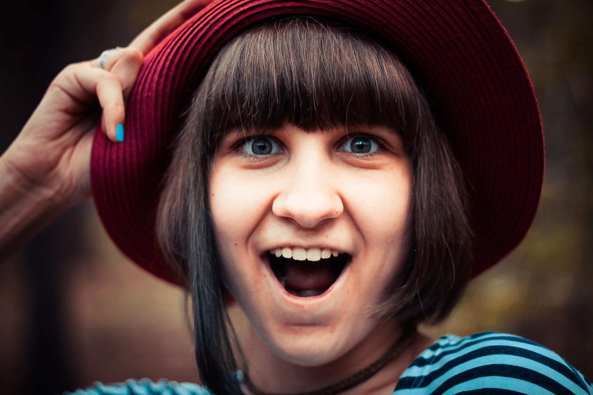 A person with short hair, wearing a wide-brimmed red hat and a striped shirt, is captured mid-expression with their mouth open, eyes wide, and one hand holding the brim of the hat. The background is blurred.