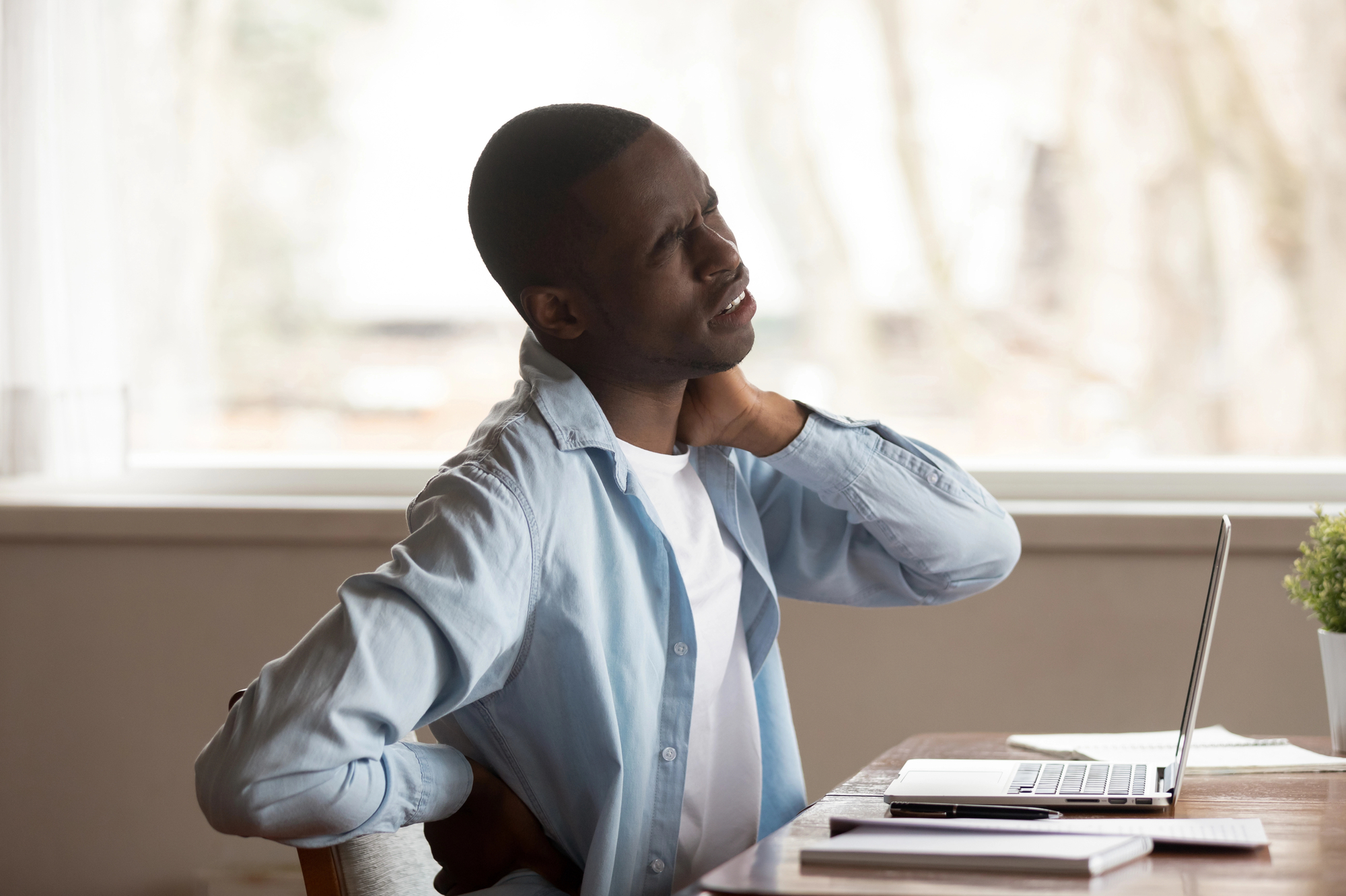 A man sitting at a desk holds his neck and back in discomfort. He is wearing a light blue shirt over a white T-shirt. In front of him are a laptop, a notebook, and a cup, with a plant on the desk. A window in the background reveals a blurred outdoor scene.