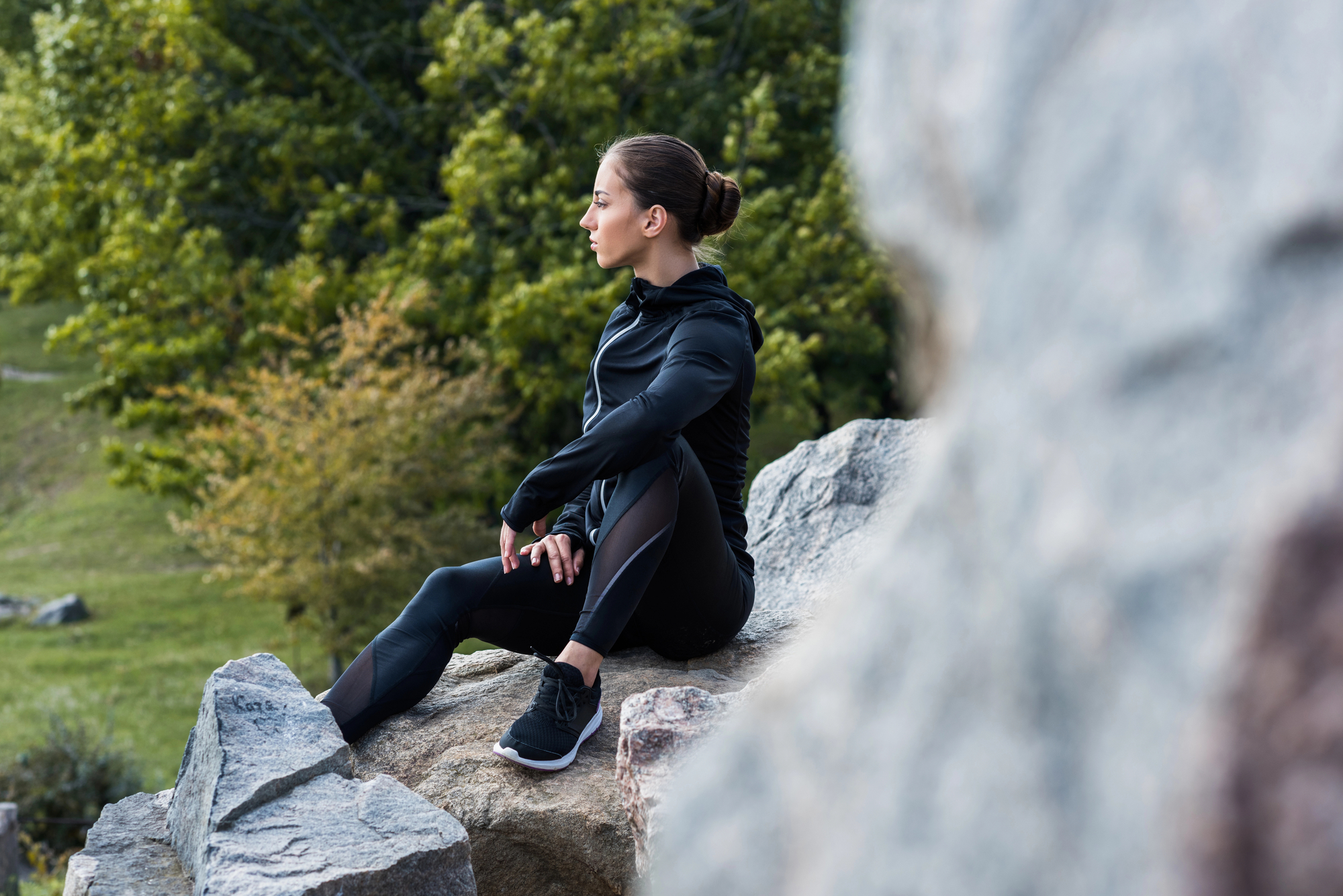 A woman in a black athletic outfit sits on a rock, gazing thoughtfully into the distance. She appears to be in a natural outdoor setting with greenery and trees in the background. She has her hair tied back in a bun and is wearing black running shoes.