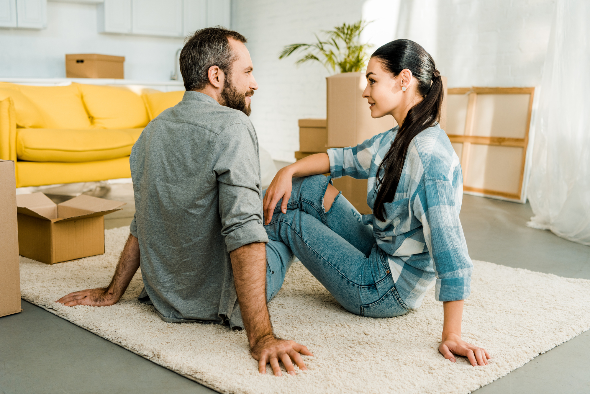 A man and a woman, both casually dressed, are sitting on a white carpet in a brightly lit room. The room has several moving boxes and a yellow couch in the background. Both individuals are smiling at each other, suggesting they are in the process of moving in together.