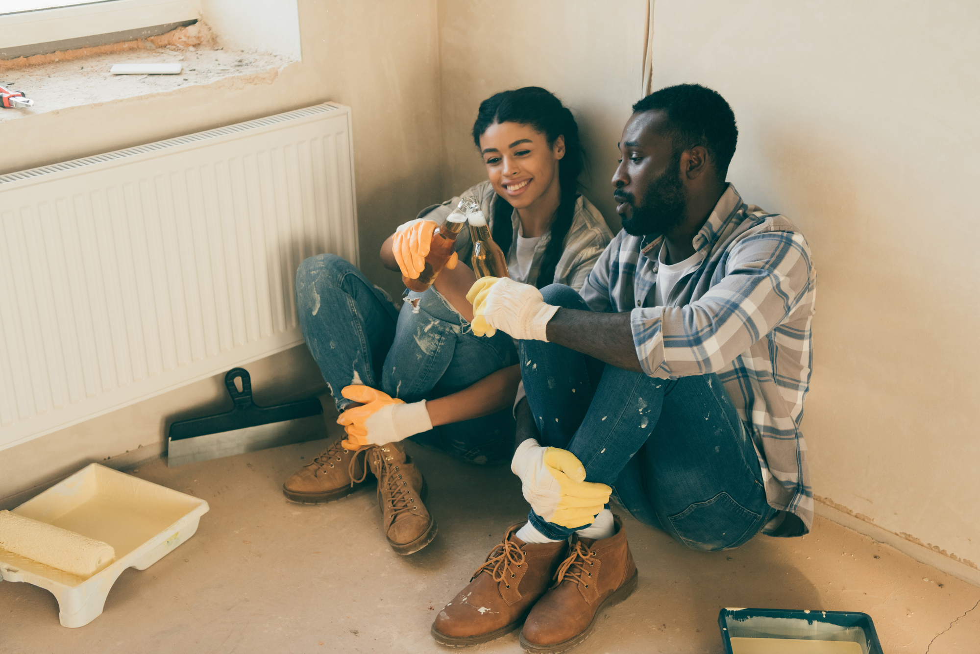 A couple takes a break while renovating, sitting on the floor by a wall. Both are wearing casual clothes and work gloves. The man is offering the woman a drink, and they both look happy and relaxed. Renovation tools and paint supplies are scattered around them.