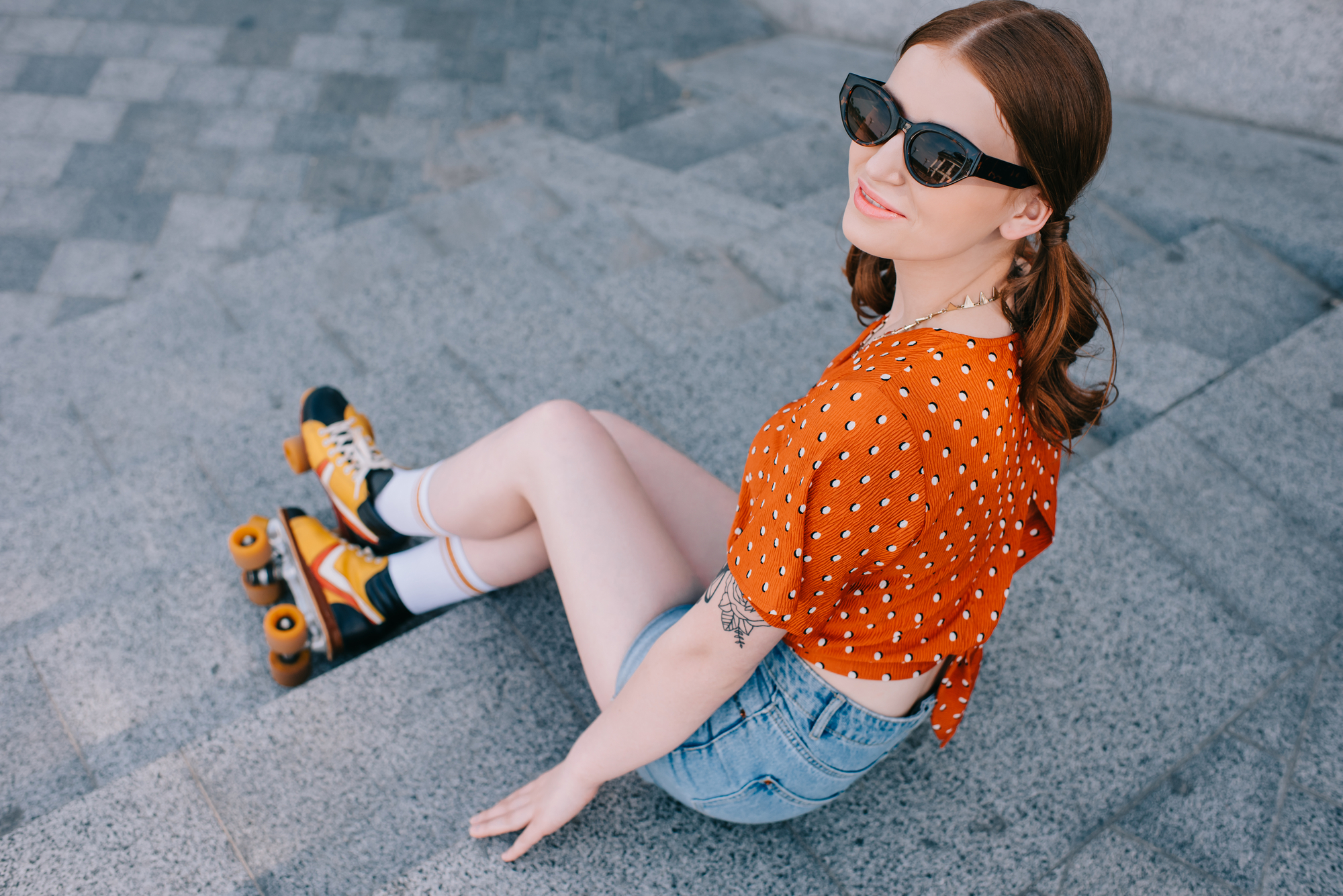A young woman wearing sunglasses, an orange polka-dot shirt, denim shorts, knee-high socks, and roller skates sits on concrete steps. She looks up and smiles at the camera, enjoying a sunny day.