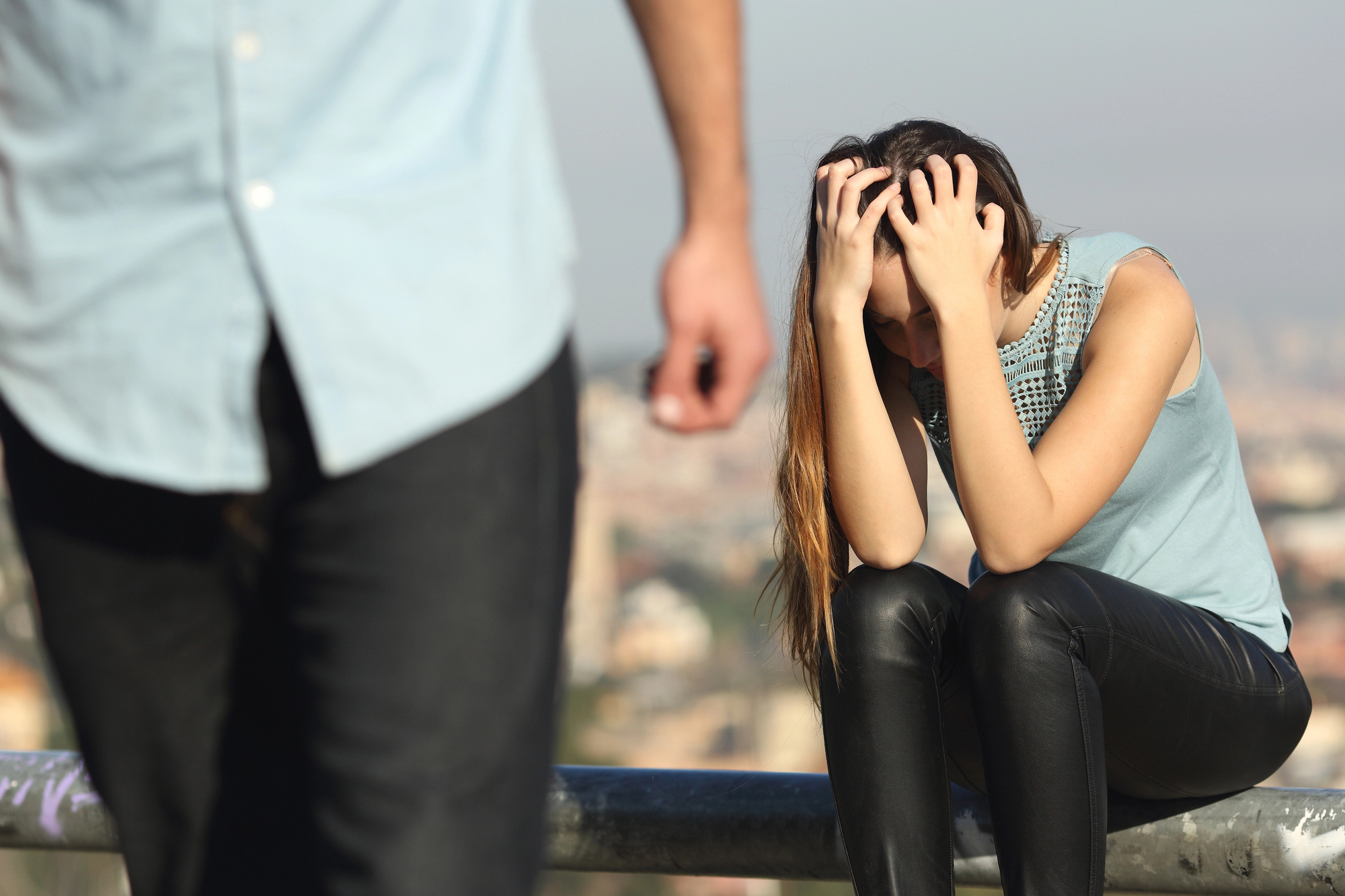 A woman sits on a rail, head in hands, appearing distressed. She is wearing a light blue sleeveless top and black pants. In the foreground, there is a person walking away, partially out of frame. The background shows a blurred cityscape.