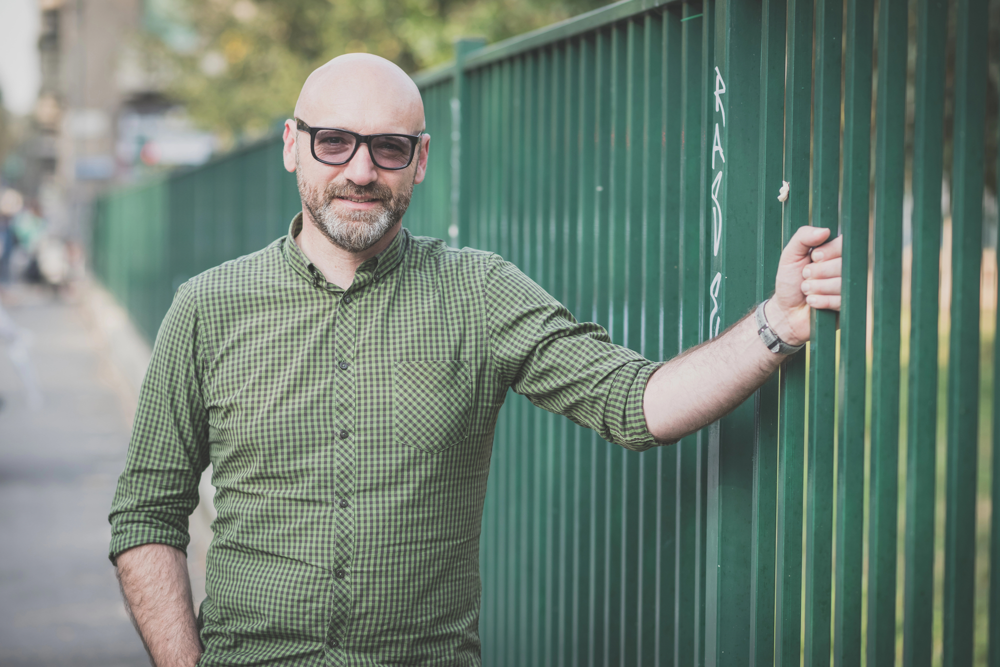A man with a bald head and glasses, dressed in a green plaid shirt, stands next to a green metal fence. He is smiling slightly and holding onto the fence with his right hand. The background is a street scene with trees and buildings.