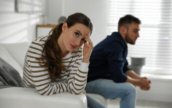 A woman with long brown hair, wearing a striped sweater, sits on a white couch looking concerned. In the background, a man with short hair, wearing a navy hoodie and jeans, sits on the edge of the couch facing away, both appearing upset in a bright room.