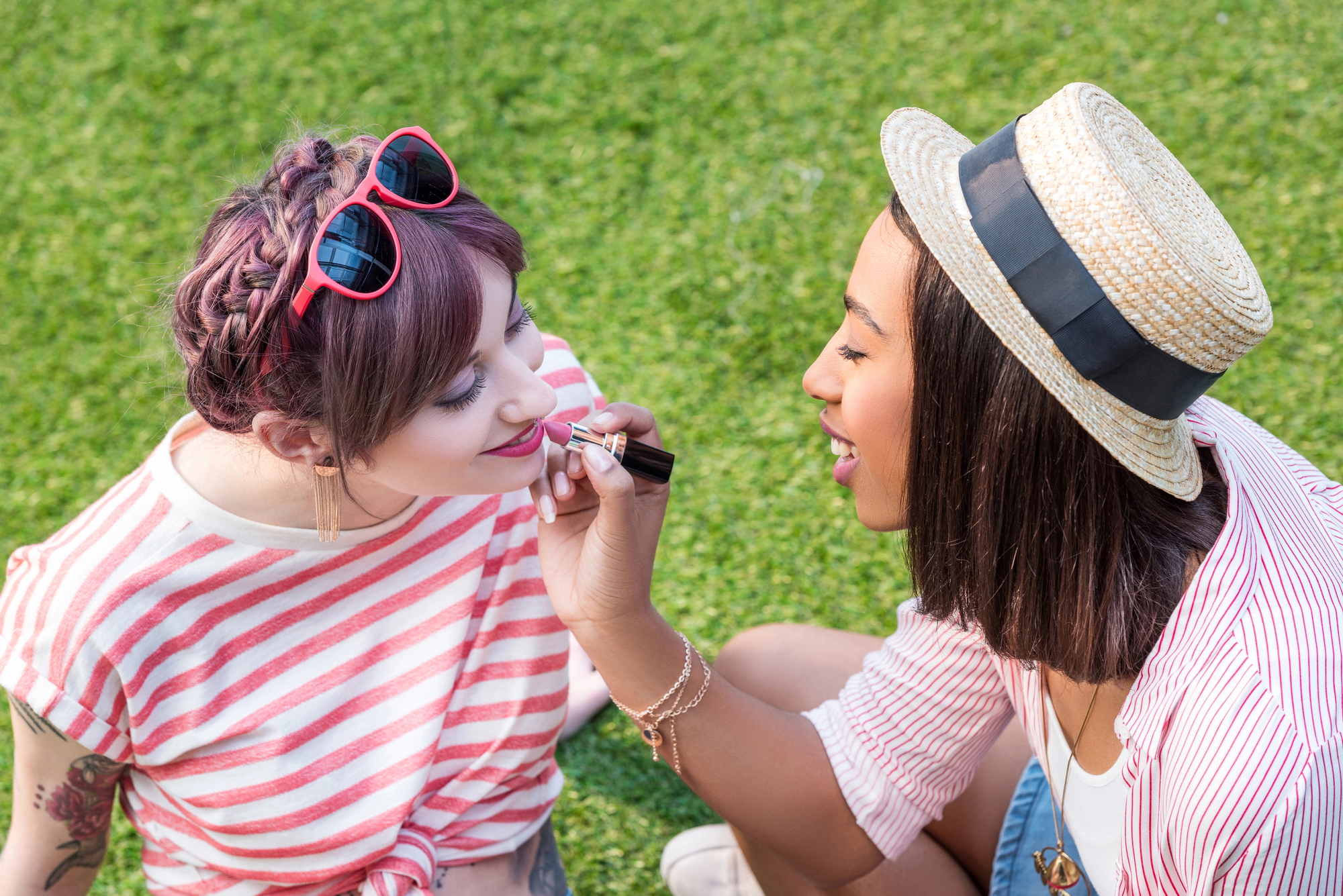 Two women are sitting on the grass. The woman on the left, wearing a striped shirt, allows her friend to apply lipstick to her lips. The woman on the right wears a hat and smiles while gently applying the lipstick. They seem to be enjoying a sunny day outdoors.
