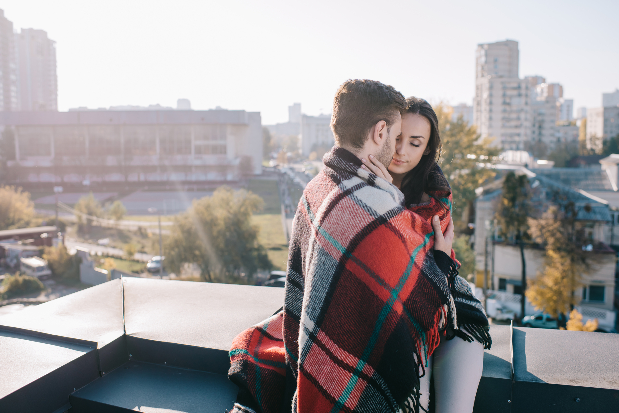 A couple sits closely together on a rooftop, wrapped in a red and black plaid blanket. The cityscape with buildings and trees is visible in the background. The sun is shining brightly, creating a serene and warm atmosphere.