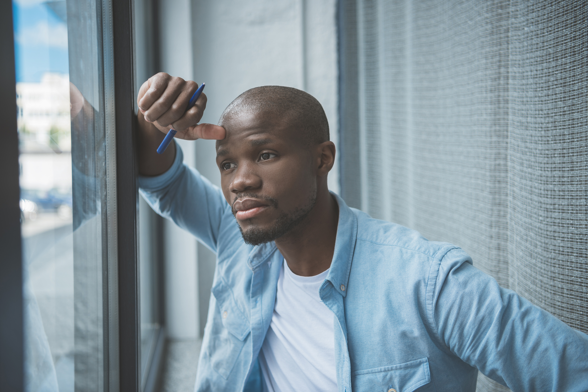 A thoughtful man gazes out of a window, resting his arm on the frame while holding a pen. He is wearing a light blue button-up shirt over a white t-shirt. The background is blurred with a partial view of the outdoors.