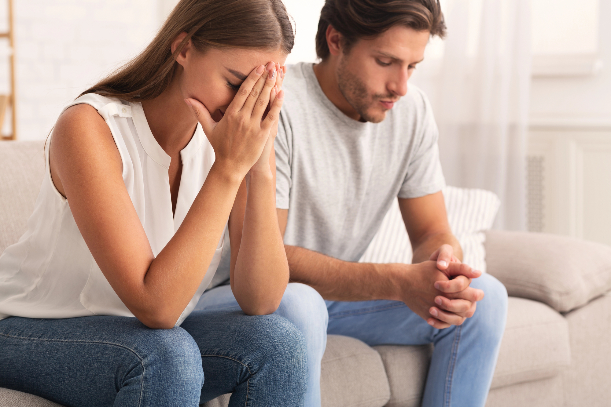 A woman sits on a couch with her head in her hands, appearing distressed. Beside her, a man sits with his hands clasped together, looking down. Both individuals seem to be in deep thought or emotion. They are in a well-lit room with a casual setting.