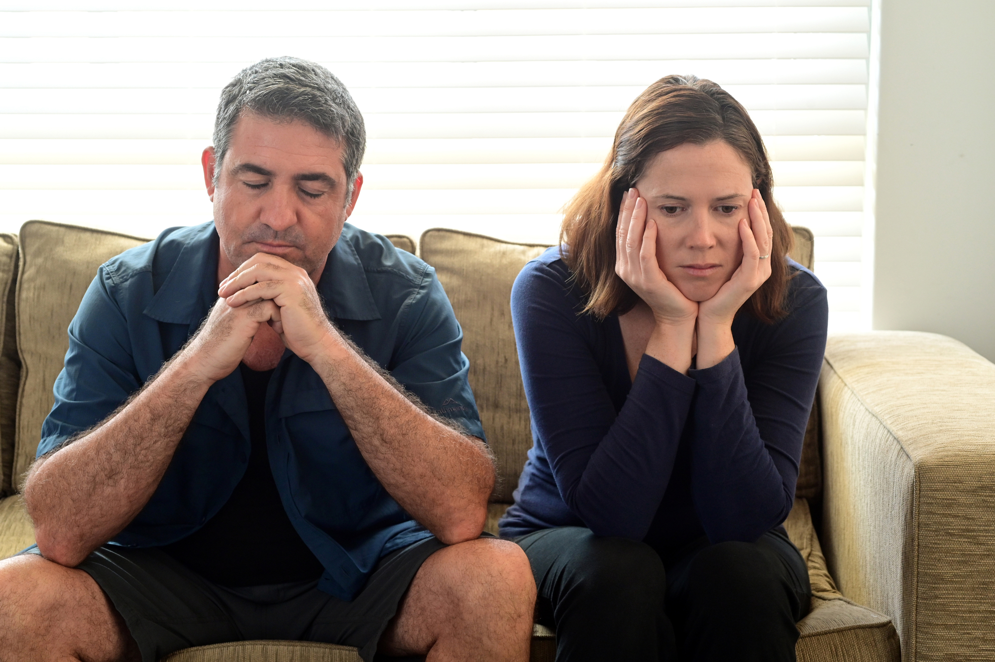 A man and a woman sit on a couch looking distressed. The man, with grey hair and a beard, has his hands clasped together and eyes closed. The woman, with shoulder-length brown hair, rests her head on her hands while looking down. Both appear deep in thought.