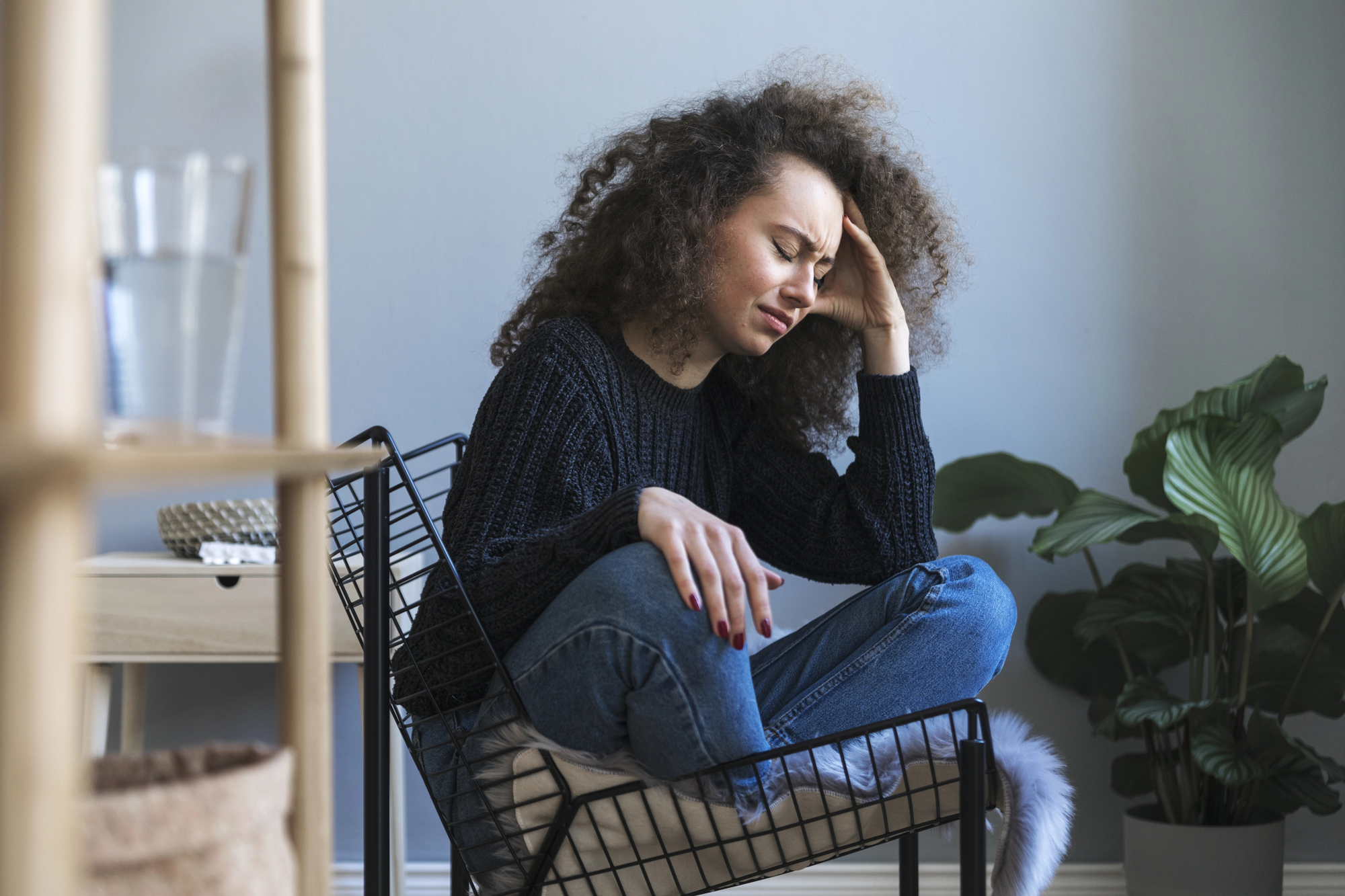A woman with curly hair sits in a modern wire chair, resting her head in one hand with a distressed expression. She is wearing a dark sweater and jeans, in a room with neutral tones and a large green plant in the corner.