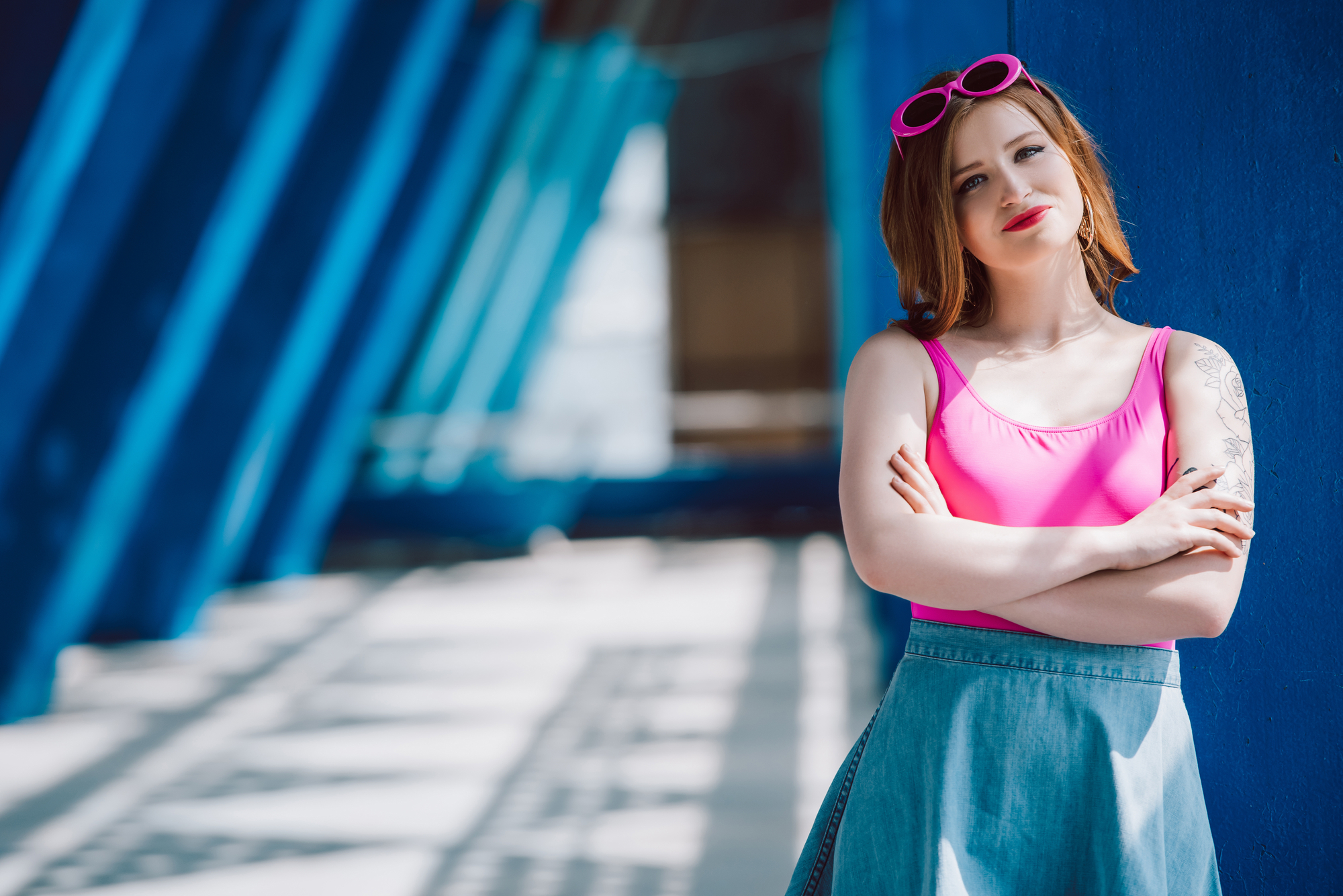 A woman with short red hair stands confidently against a blue wall. She wears a bright pink top, denim skirt, and matching pink sunglasses on her head. The background features a series of blue structural beams and patterns, with sunlight casting shadows.