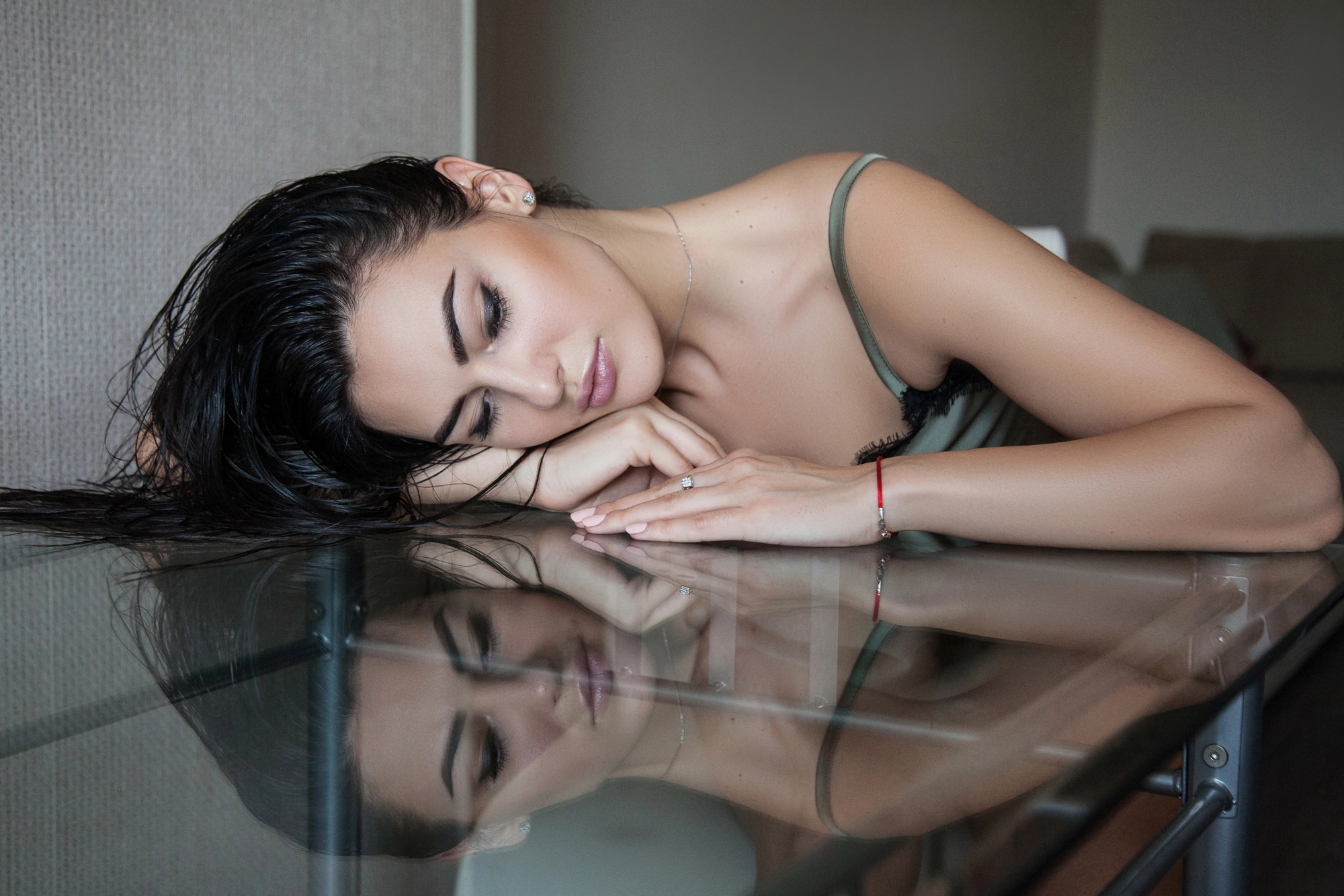 A woman with dark, wet hair is lying on her side on a glass table, her eyes closed and face resting on her crossed arms. She is wearing a green tank top and a red bracelet on her left wrist. Her reflection is visible on the surface of the table.