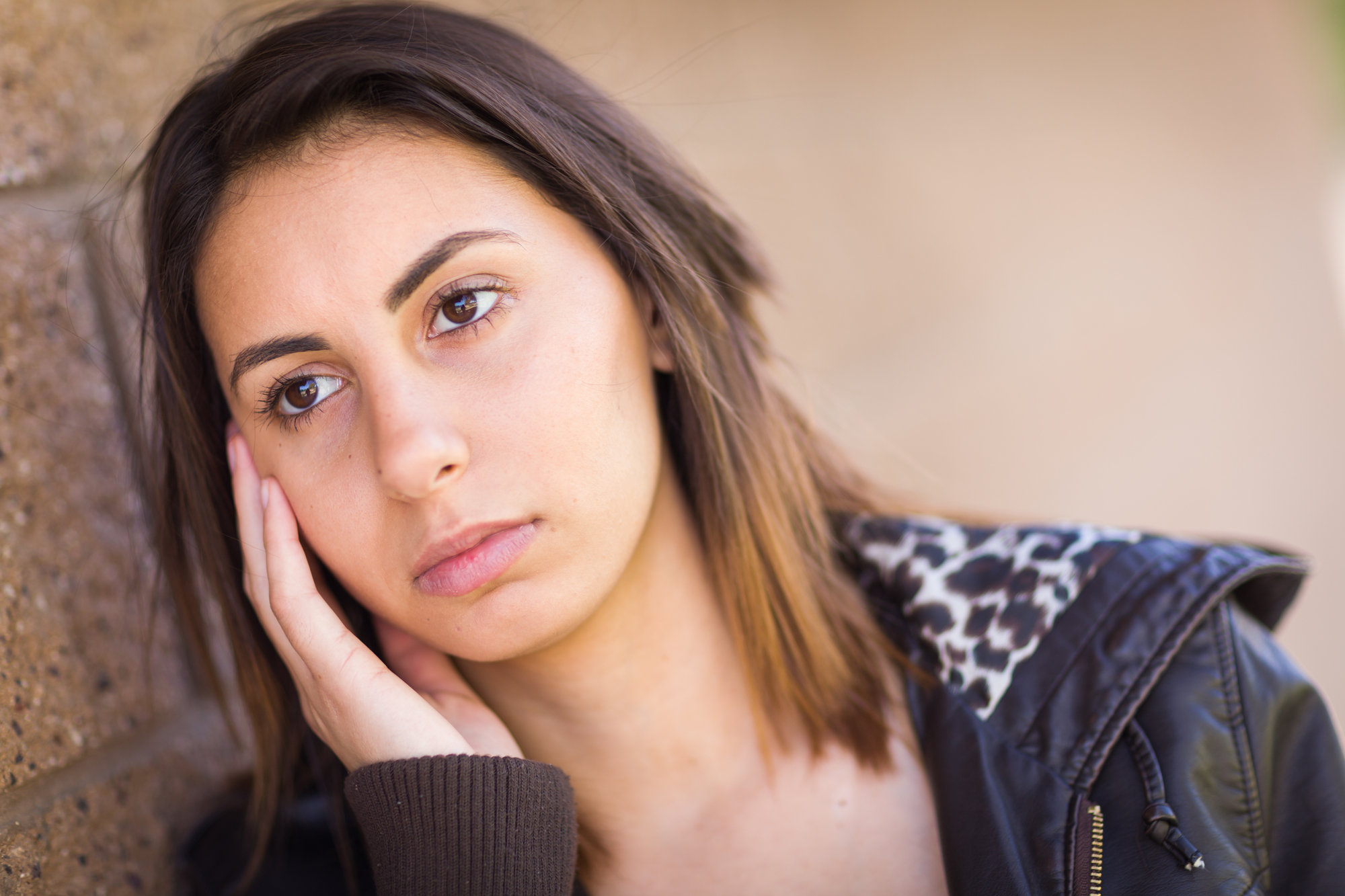 A young woman with shoulder-length brown hair leans against a wall, resting her cheek on her hand. She wears a dark jacket with a leopard print lining. She has a thoughtful or slightly pensive expression on her face.