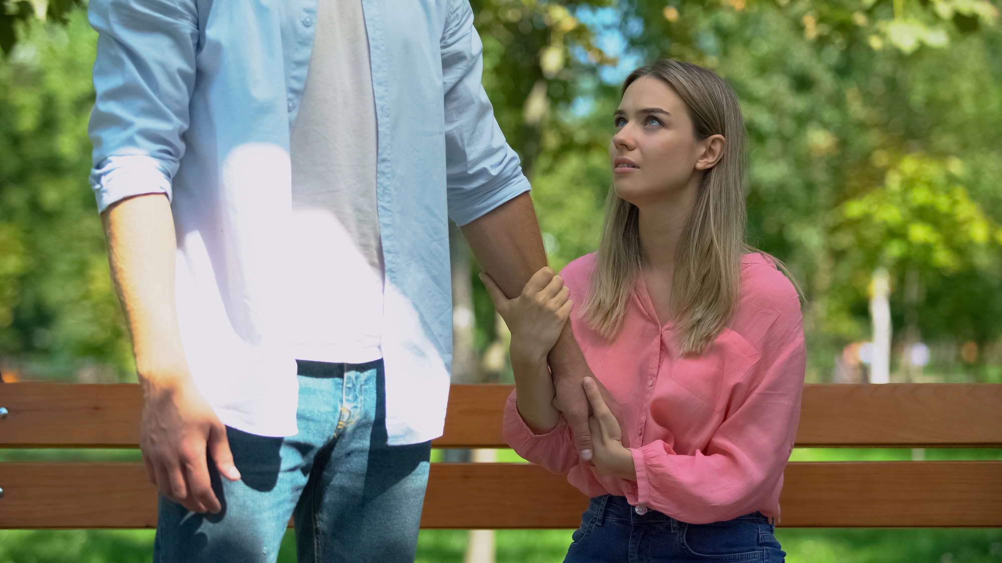 A woman in a pink blouse sits on a bench, holding onto the arm of a standing man in a blue shirt and white t-shirt. She is looking up at him with a concerned expression. The background includes trees and greenery, indicating a park setting.