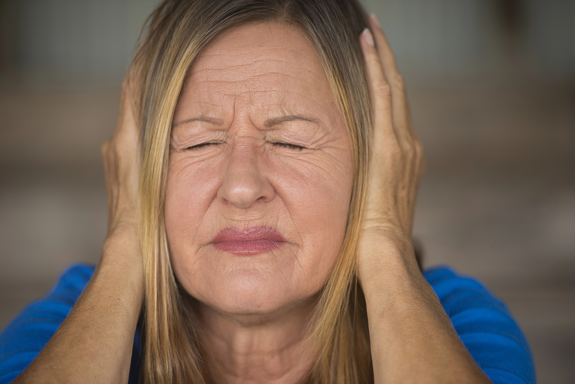 A woman with shoulder-length hair and a blue shirt closes her eyes tightly and covers her ears with her hands, appearing distressed or overwhelmed. Wrinkles are visible on her face, and she has a pained expression.
