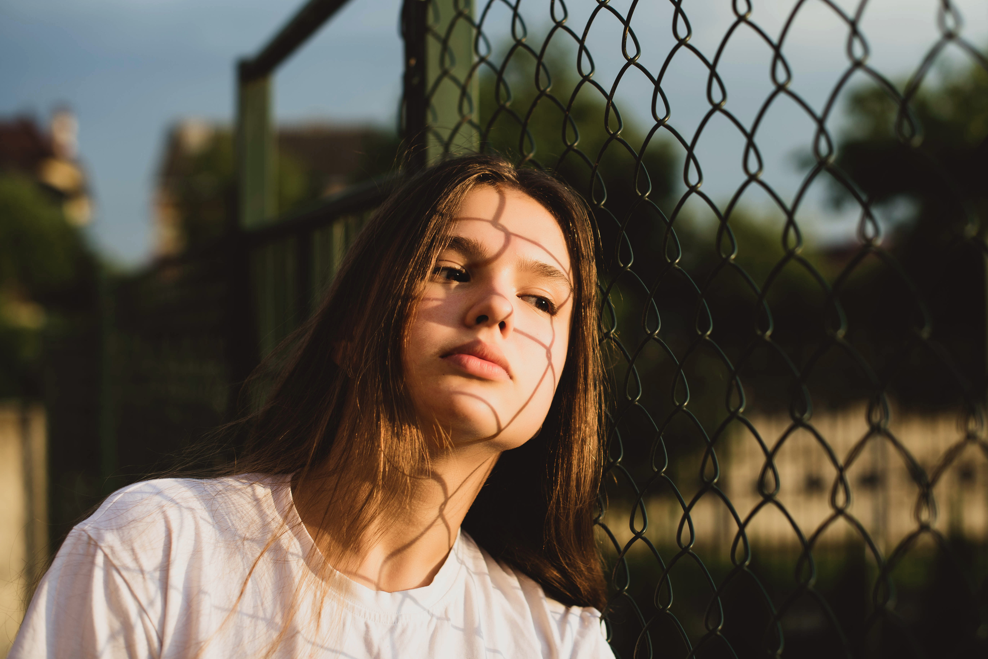 A young person with long brown hair leans against a chain-link fence, with the sun casting intricate shadow patterns on their face. They have a relaxed expression and are wearing a white T-shirt. The background is a blurred outdoor setting.