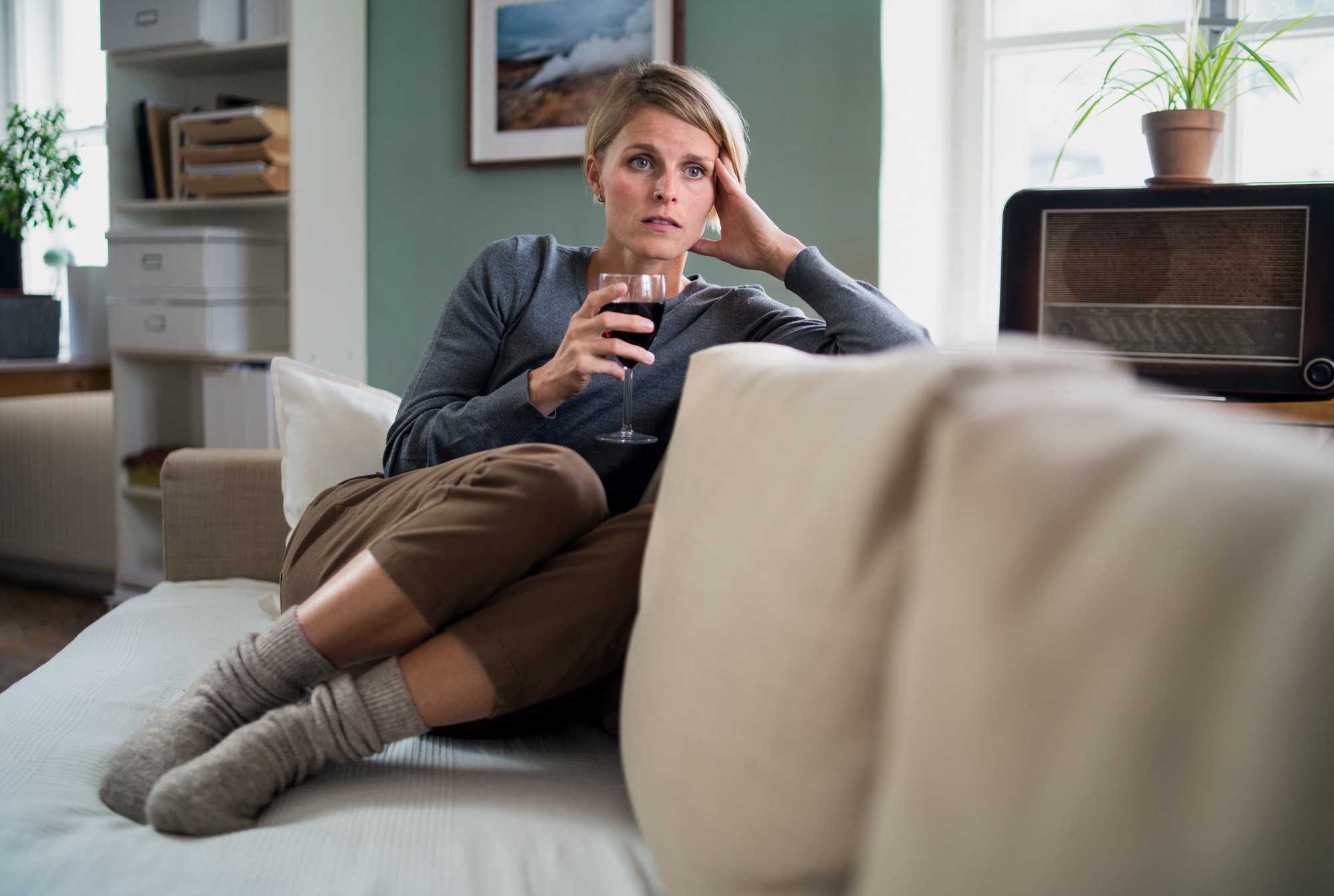 A person with short hair sits on a beige couch, holding a glass of red wine, appearing thoughtful. They wear a gray sweater, brown pants, and wool socks. The room is softly lit, featuring plants, a vintage radio, and a framed photograph on the wall.