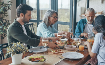 Four people are seated around a table, enjoying a meal together. The table is set with various dishes, including a salad, bread, and other foods. The setting appears to be a home with large windows in the background, allowing natural light to illuminate the scene.