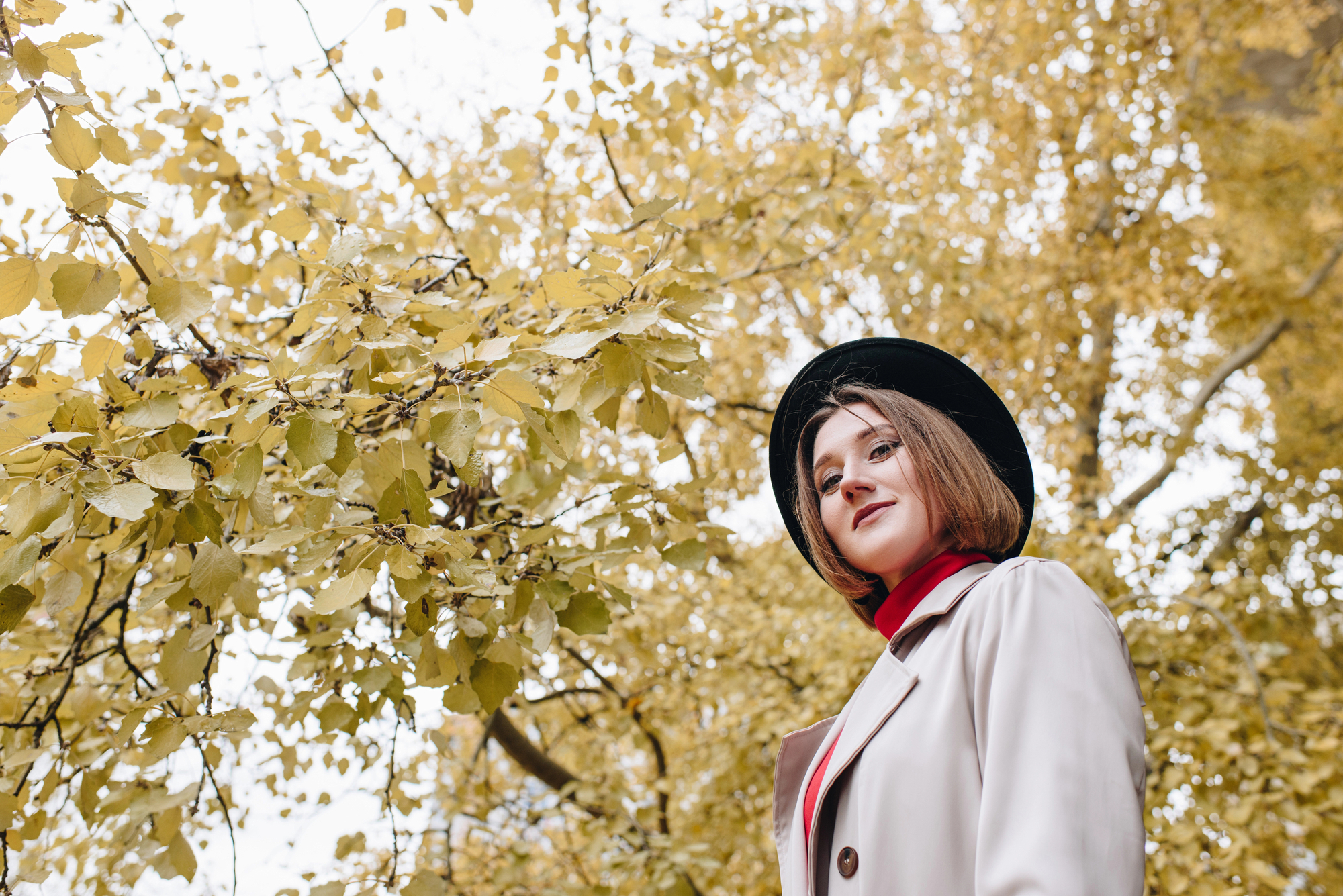 A person with short hair is wearing a black hat and beige coat, standing near a tree with yellow leaves. They look content, with their face turned slightly to the side. The autumn foliage creates a vibrant backdrop against the pale sky.