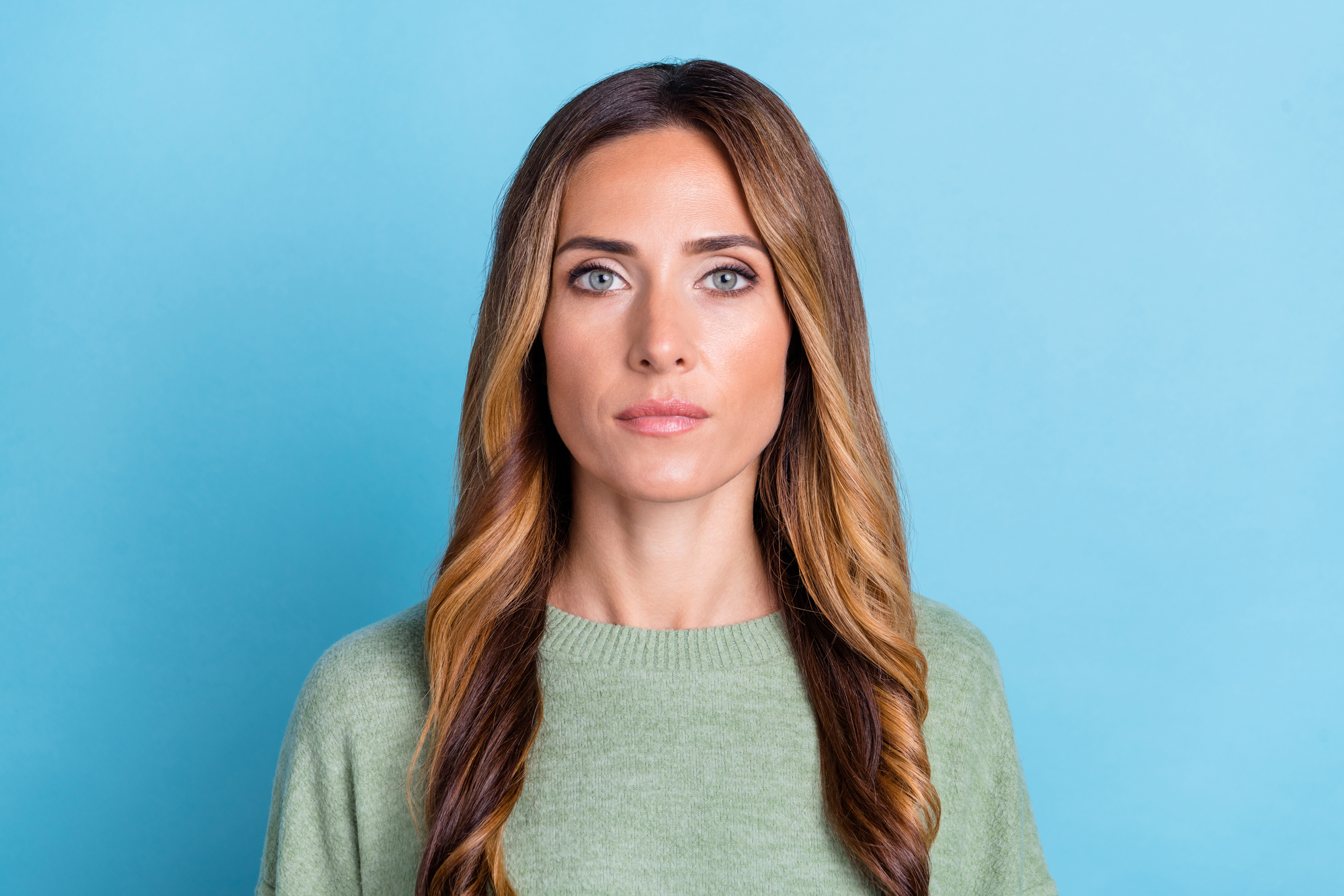 A woman with long, wavy brown hair featuring blonde highlights is wearing a light green sweater and standing against a light blue background. She has a serious expression and is looking directly at the camera.