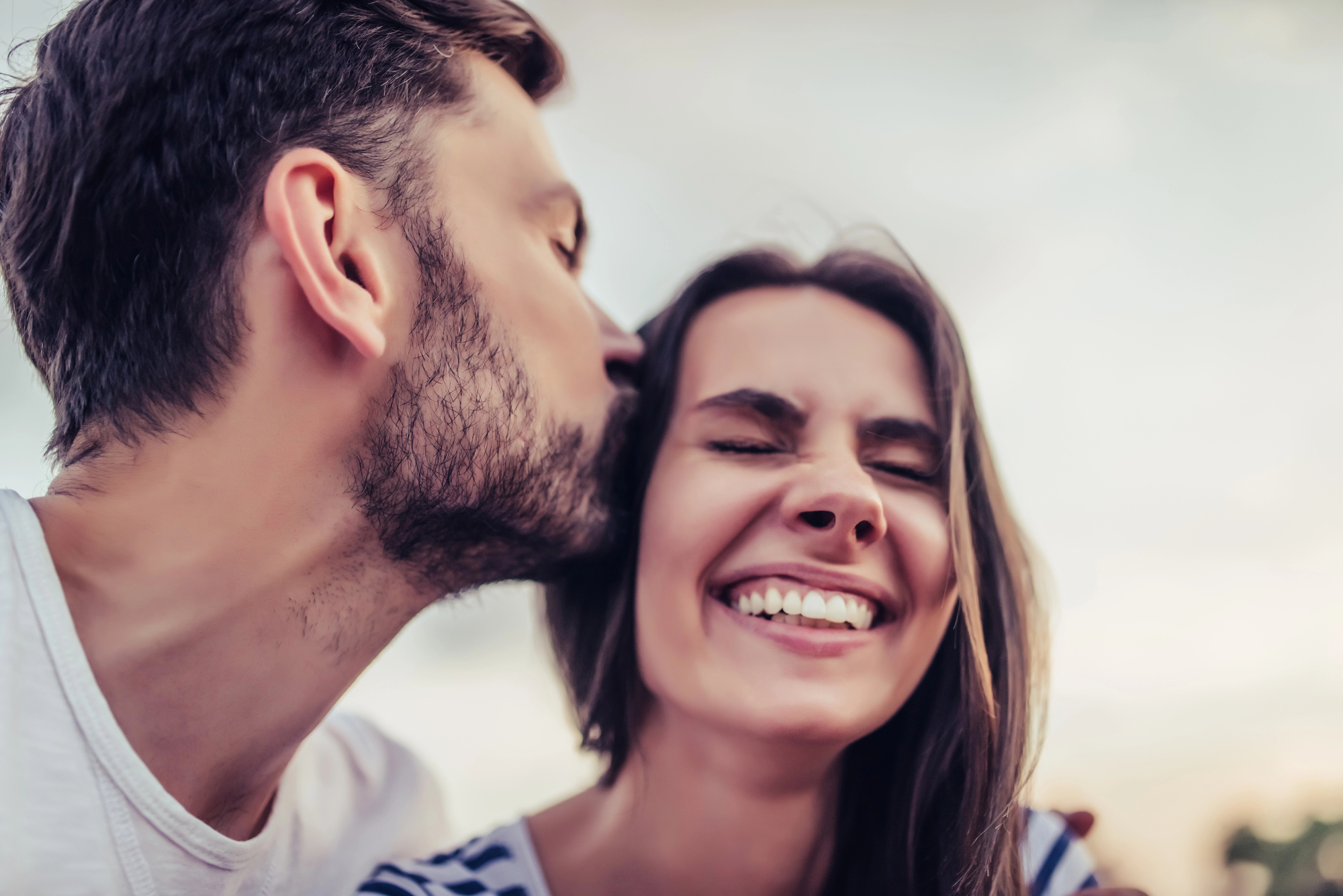 A man with a beard kisses a smiling woman on the cheek. The woman has long, brown hair and her eyes are closed in happiness. They are outdoors and appear to be enjoying a pleasant, affectionate moment together.