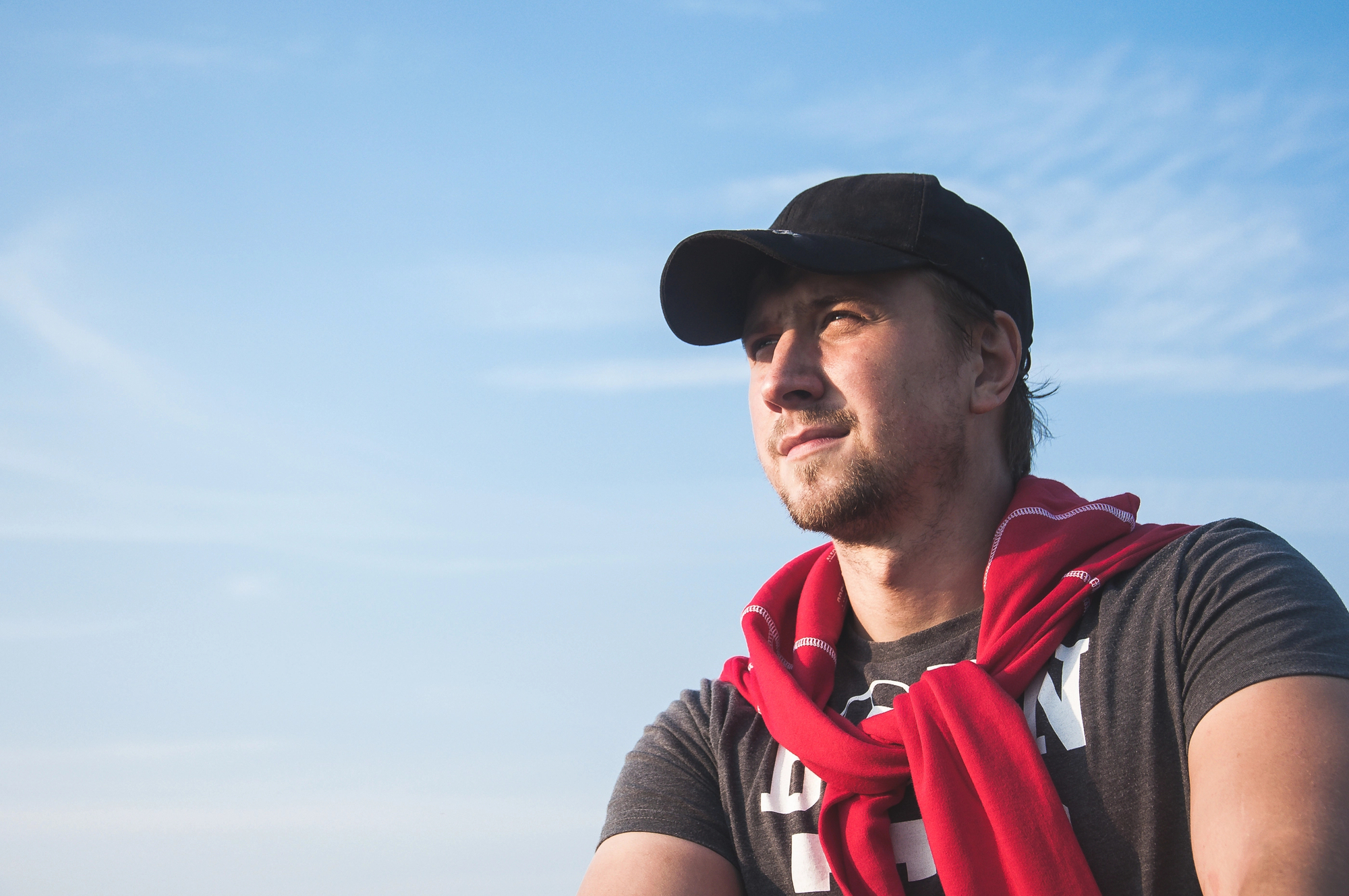A man wearing a black cap and a red scarf draped over his shoulders gazes to his left against a clear blue sky background. He has a light beard and is dressed in a dark t-shirt. The bright sky provides a serene backdrop to his contemplative expression.