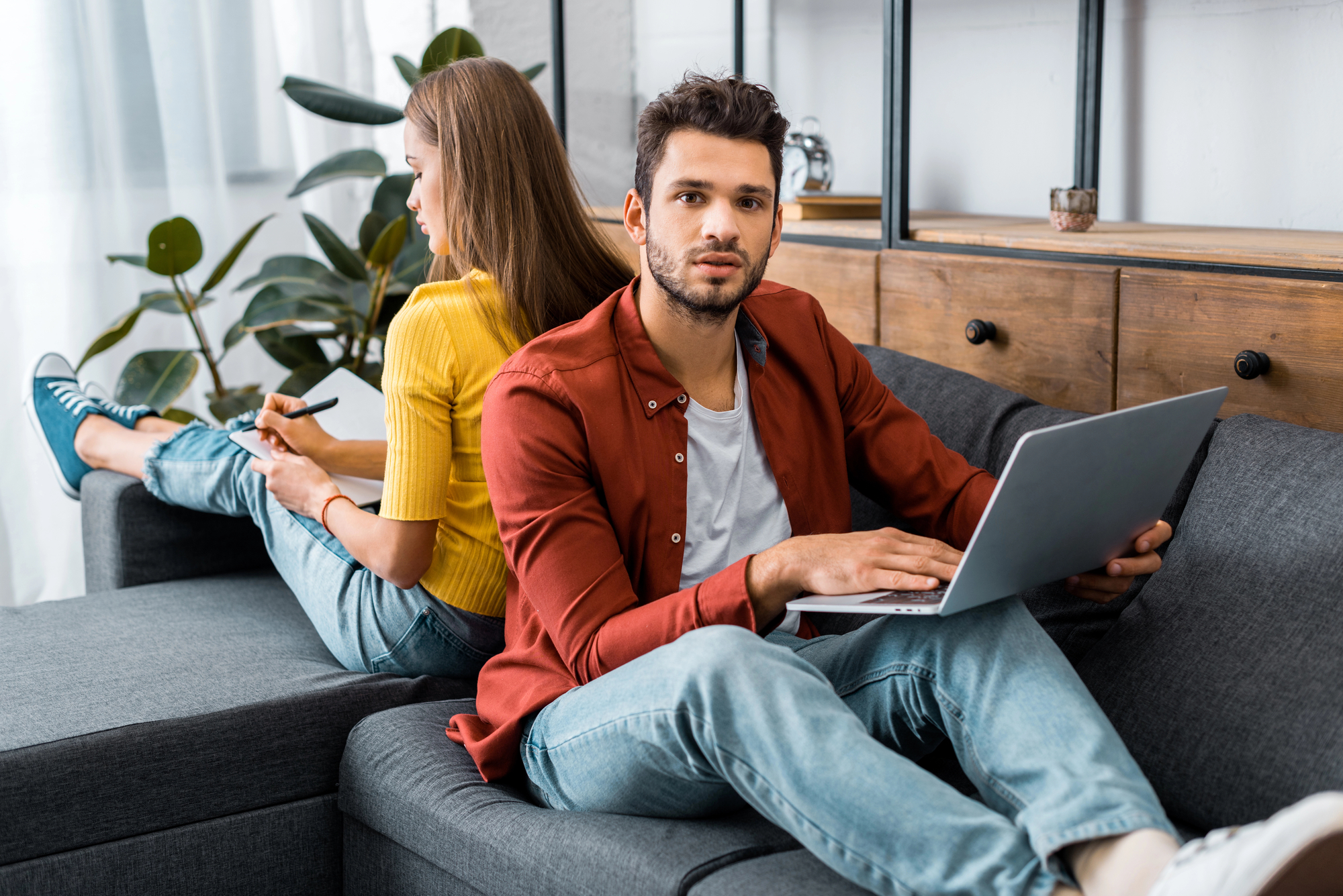 A man and woman sit back-to-back on a sofa. The man, wearing a red shirt and jeans, uses a laptop on his lap. The woman, in a yellow shirt and jeans, writes in a notebook. They are in a modern living room with plants and wooden furniture in the background.