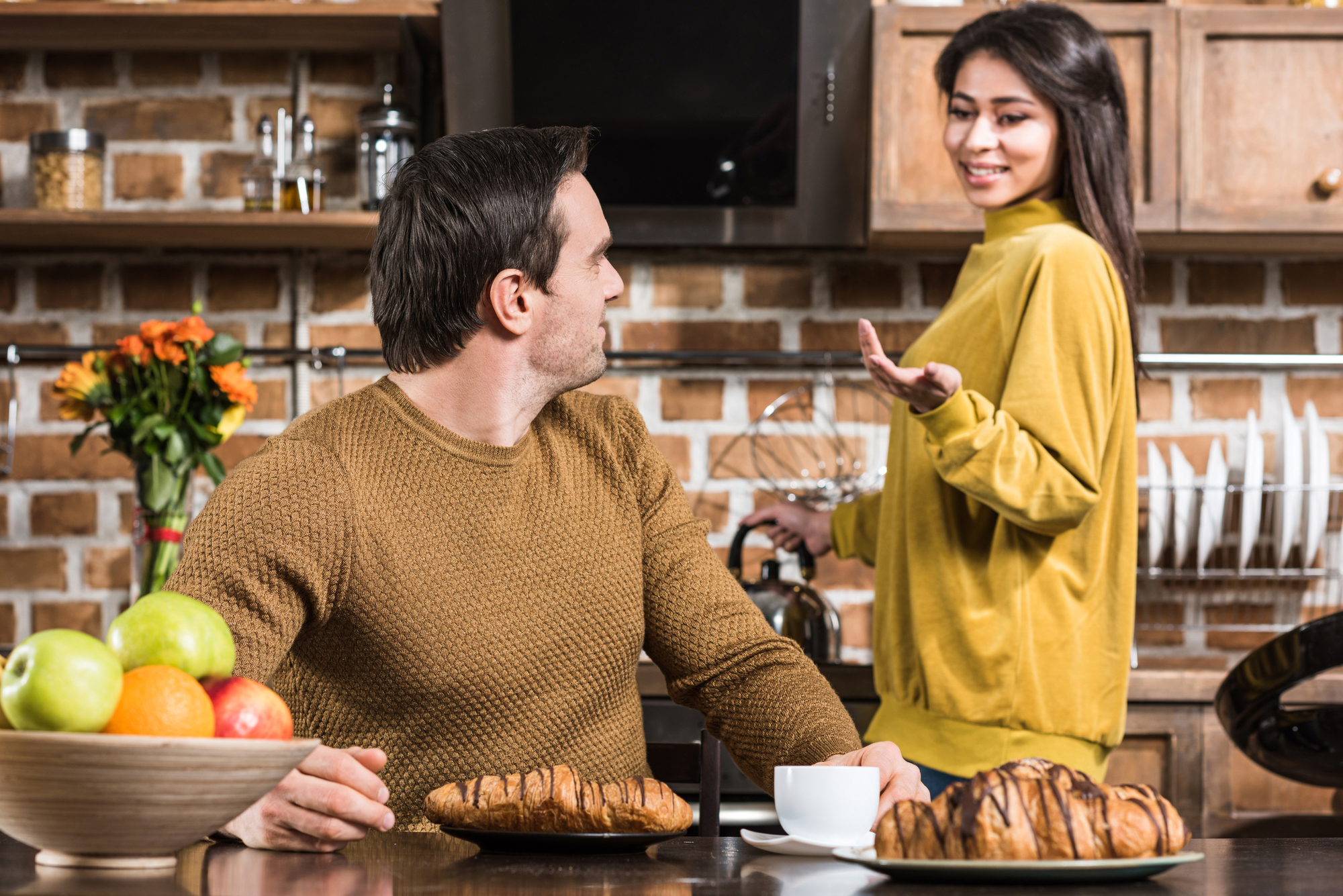 A man and woman are in a kitchen, conversing. The man is seated at a table with a cup of coffee and pastries, looking at the woman. The woman stands, gesturing with one hand while holding a coffee pot. The kitchen features brick walls and wooden cabinets.