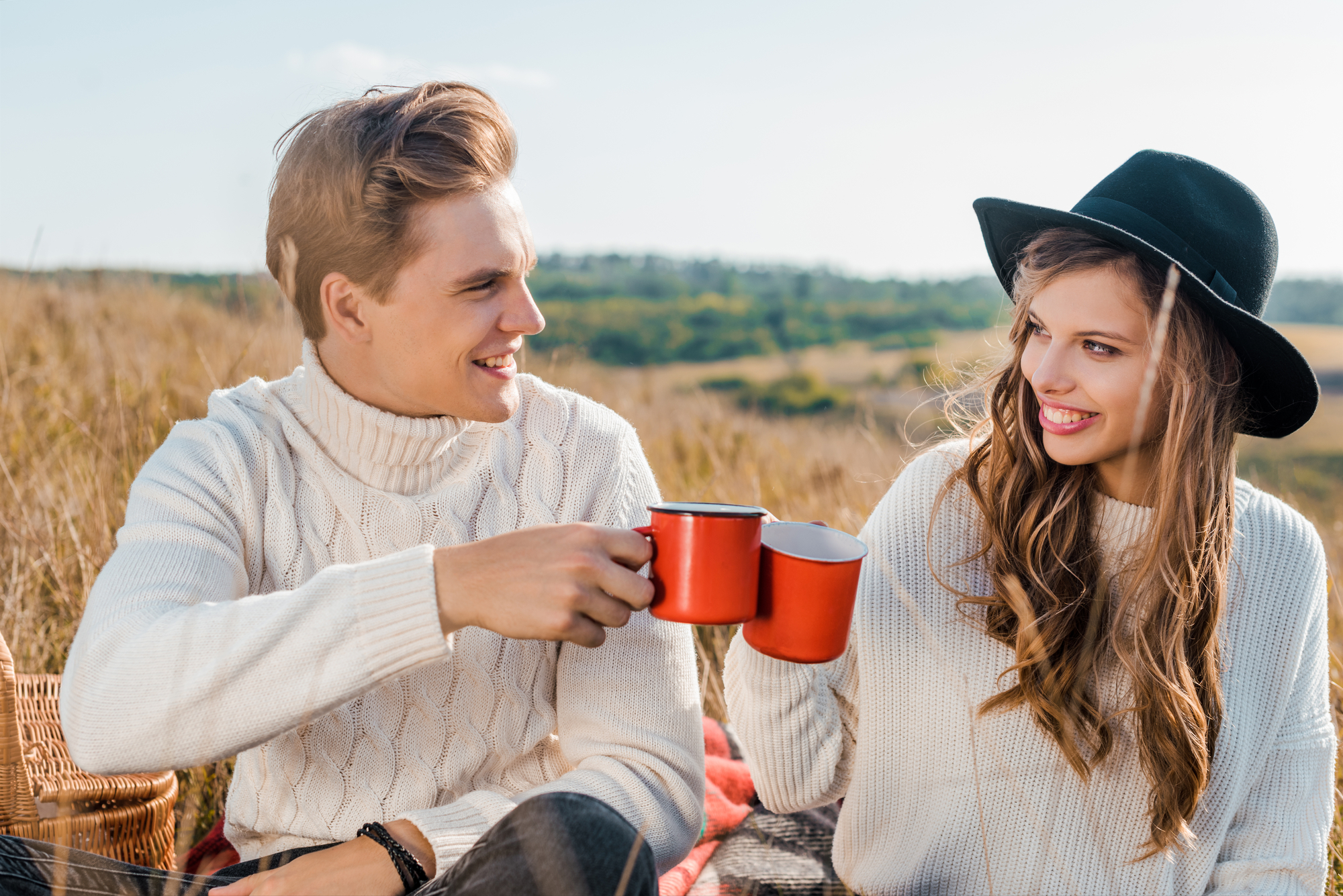 A man and a woman, both wearing white sweaters, are sitting outdoors on a blanket, toasting with red mugs. The woman is wearing a black hat, and they are smiling at each other. There is a wicker basket beside them, and a scenic landscape in the background.