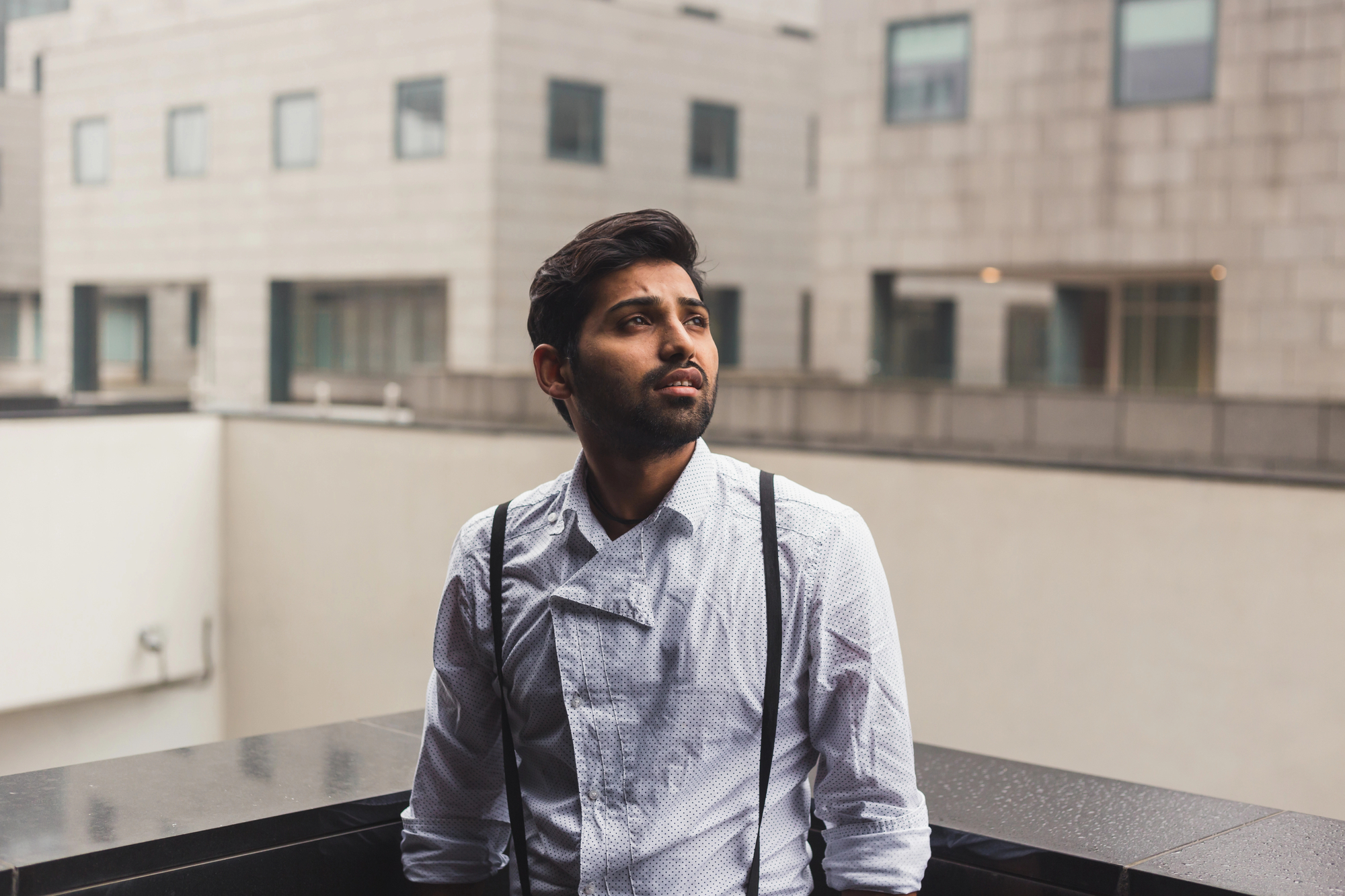 A man with a beard wearing a white shirt and black suspenders stands outdoors, leaning on a black railing. He gazes thoughtfully into the distance. The background features modern buildings with large windows and a neutral color palette.