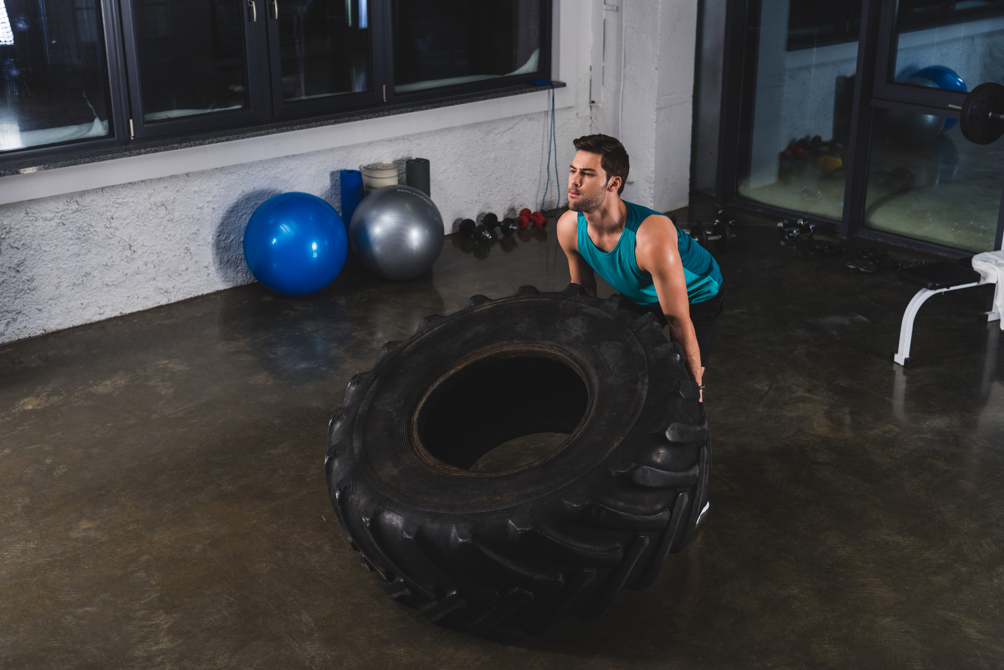 A man in a blue tank top is lifting a large tire in a gym. He appears focused and determined. There are exercise balls and other gym equipment in the background. The floor is polished concrete.