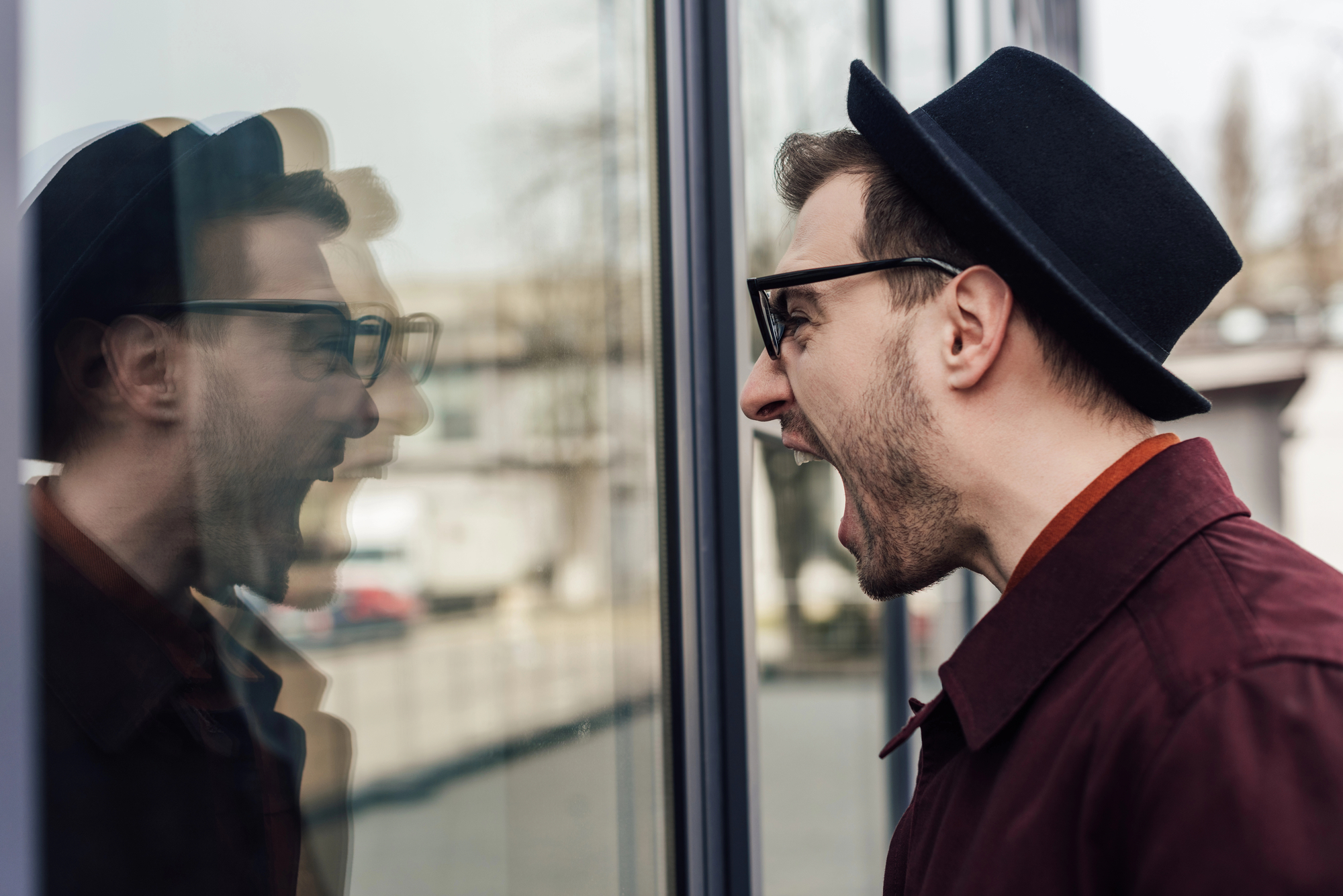 A man wearing a black hat and glasses is shown yelling at his reflection in a glass window. The background shows an outdoor setting, slightly blurred. The man is wearing a maroon jacket and appears to be expressing frustration or anger.
