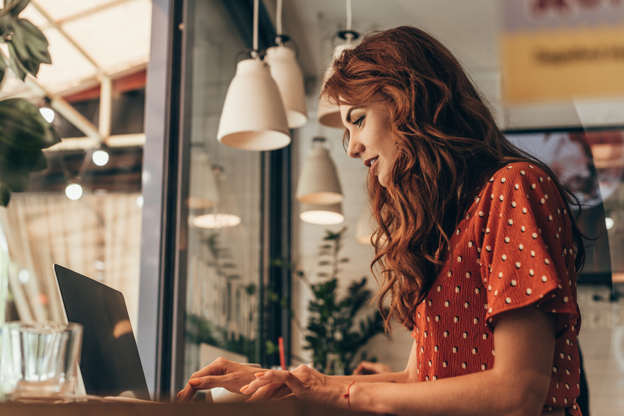 A woman with long red hair types on a laptop at a cozy café. She is wearing a red polka-dotted dress and appears focused on her work. Pendant lights hang above her, and there are plants and a glass of water nearby. The atmosphere is warm and inviting.