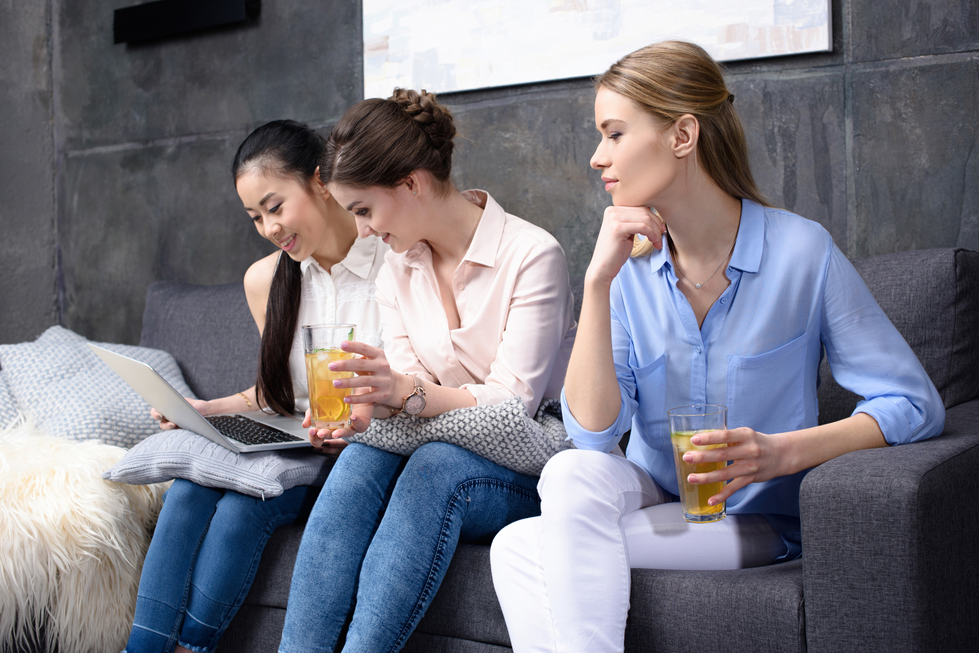 Three women are sitting on a gray couch. Two of them, wearing blue jeans and light-colored tops, are looking at a laptop screen and sharing a laugh while holding drinks. The third woman, in white pants and a blue shirt, holds a drink and looks towards them, smiling.