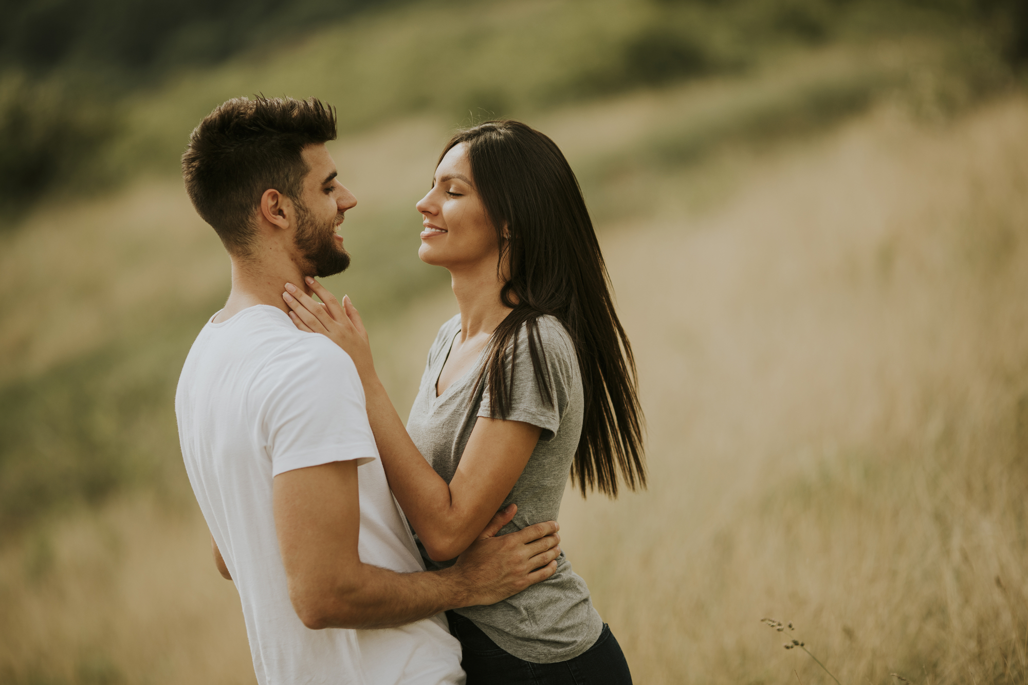 A couple stands in a grassy field, facing each other and smiling. The man, wearing a white t-shirt, has his arm around the woman, who has long dark hair and is wearing a gray t-shirt. The background is blurred, highlighting the couple in the foreground.