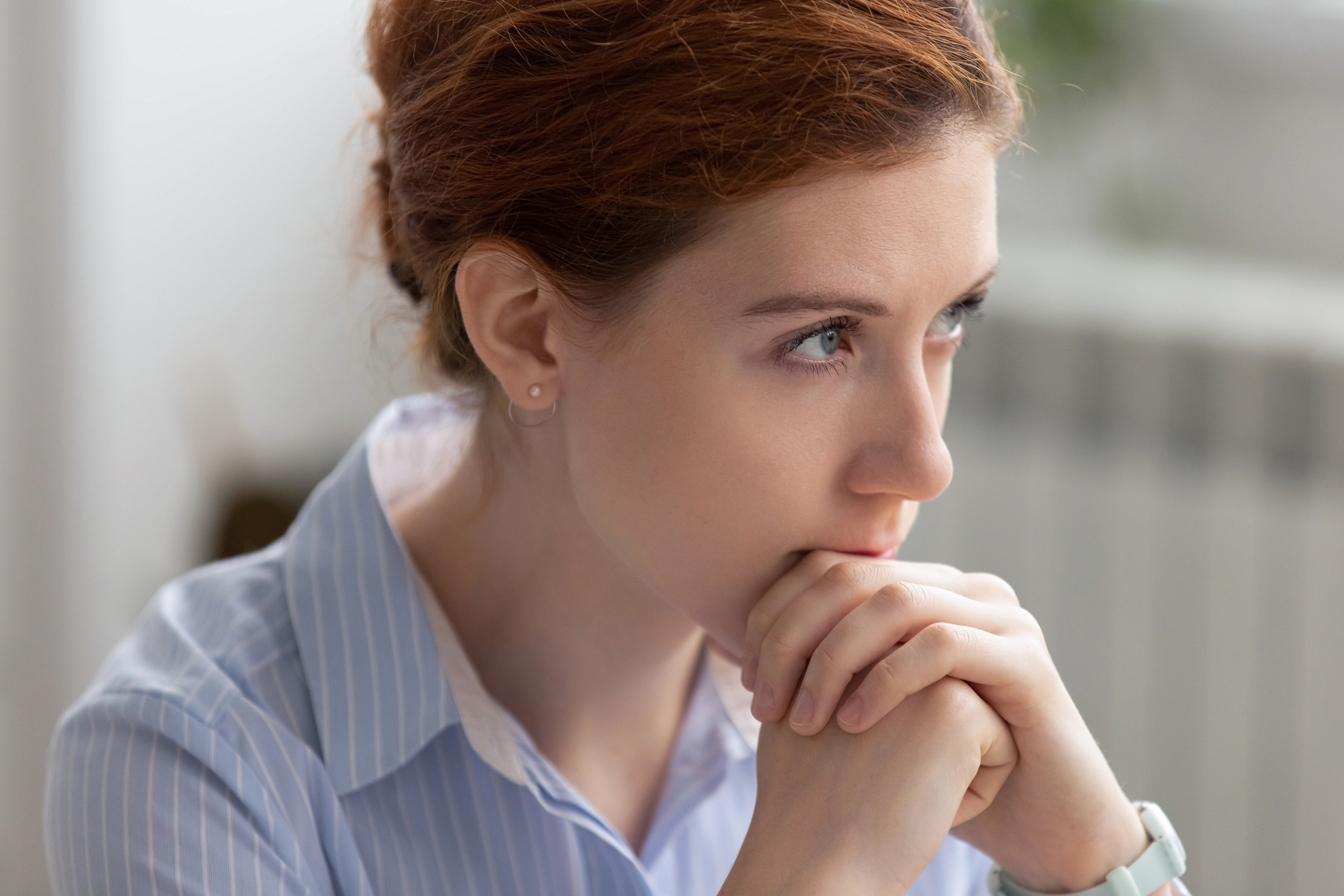 A person with red hair, wearing a light blue striped shirt, is seated indoors. The individual is resting their chin on clasped hands, appearing deep in thought and facing slightly to the right. The background is softly blurred, indicating an indoor setting.