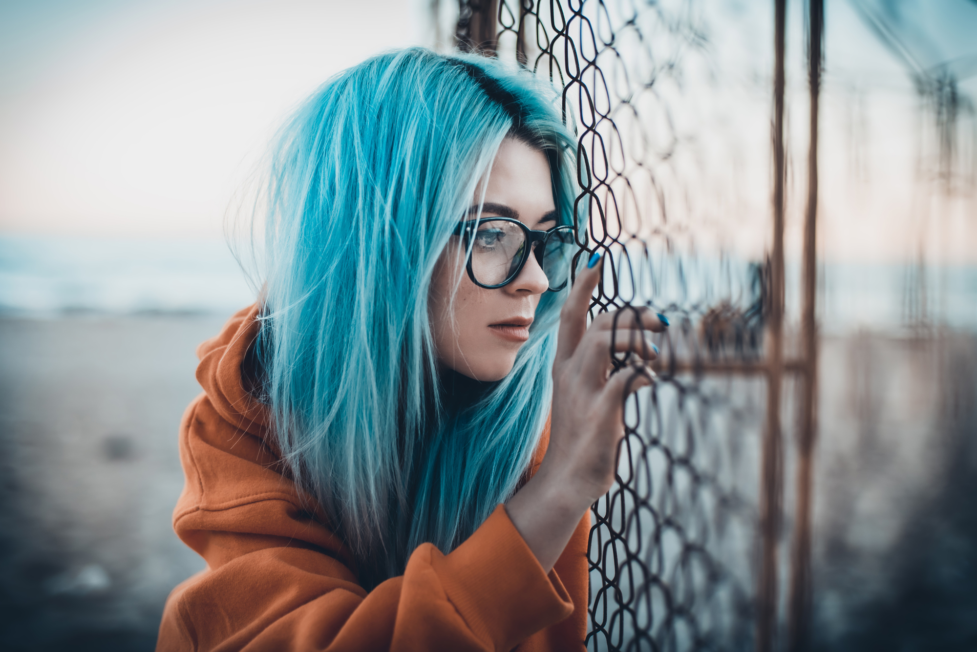 A person with long, vibrant blue hair and glasses is standing beside a wire fence, gazing thoughtfully through it. They are wearing an orange hoodie and appear to be near a beach,with the sea and sky visible in the background.
