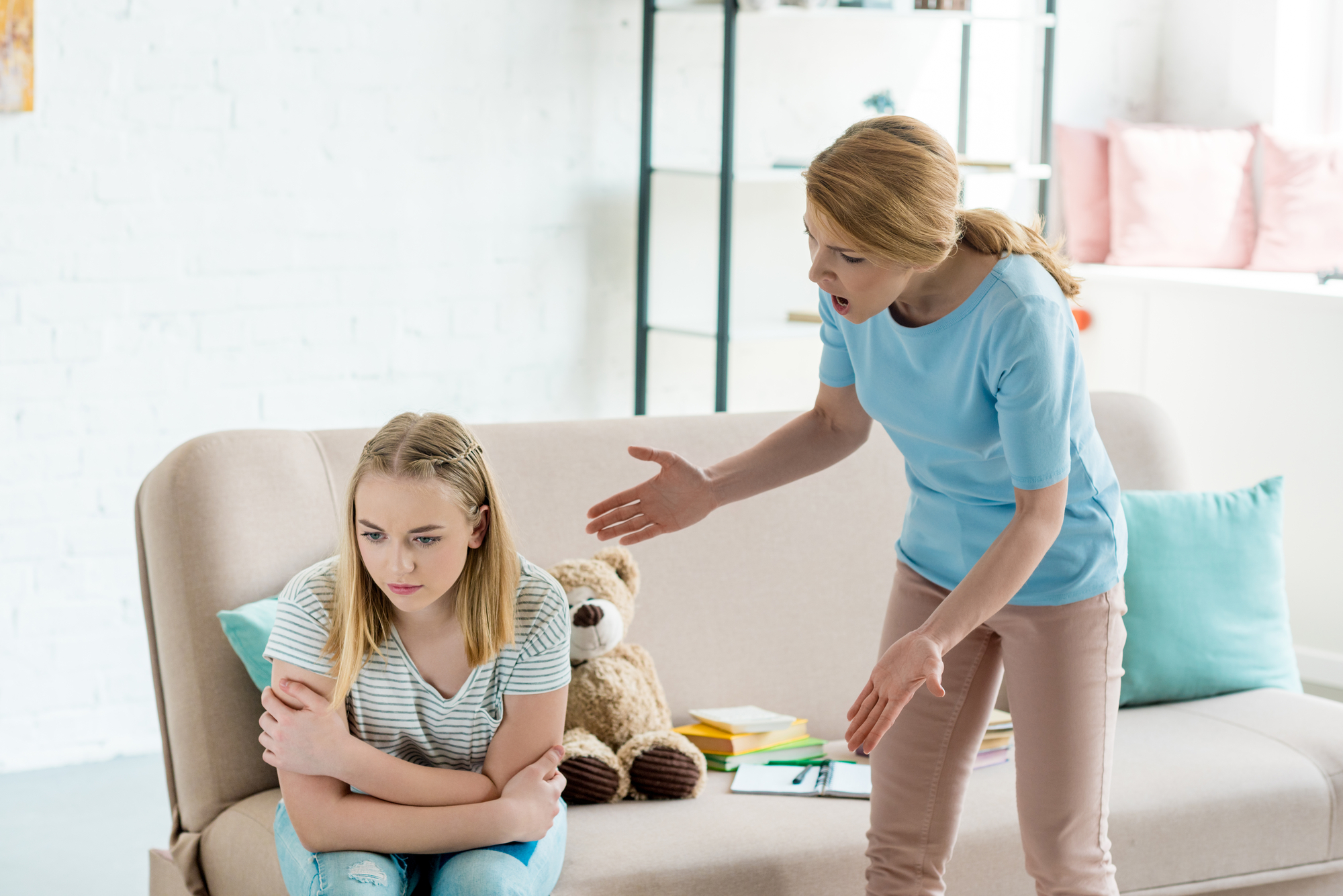 A woman is shown speaking angrily to a young girl sitting on a couch. The girl, with blonde hair, appears upset and is looking down while hugging her knees. Behind them is a stuffed teddy bear and books. The room is bright with natural light from a nearby window.